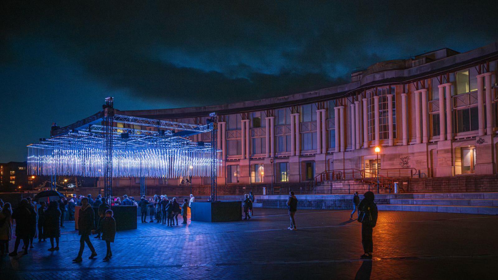 A light installation is next to Bristol's amphitheatre. Lights are hanging down from a metal structure and people are walking past it at night during the Bristol Light Festival