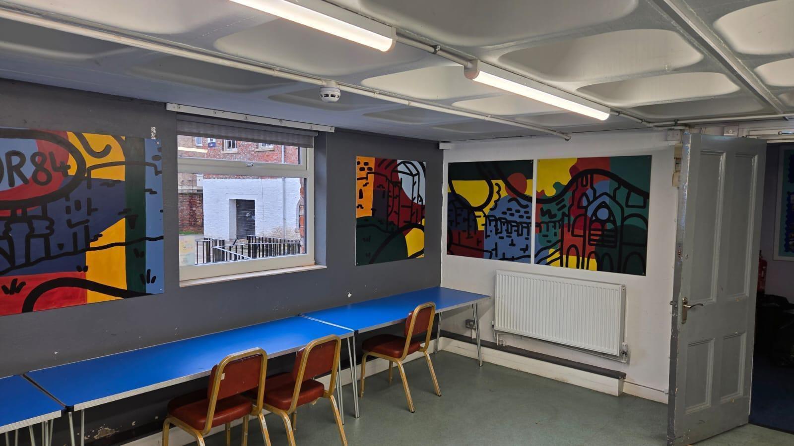 Community centre room with grey and white walls, blue tables and four colourful abstract murals on the walls.