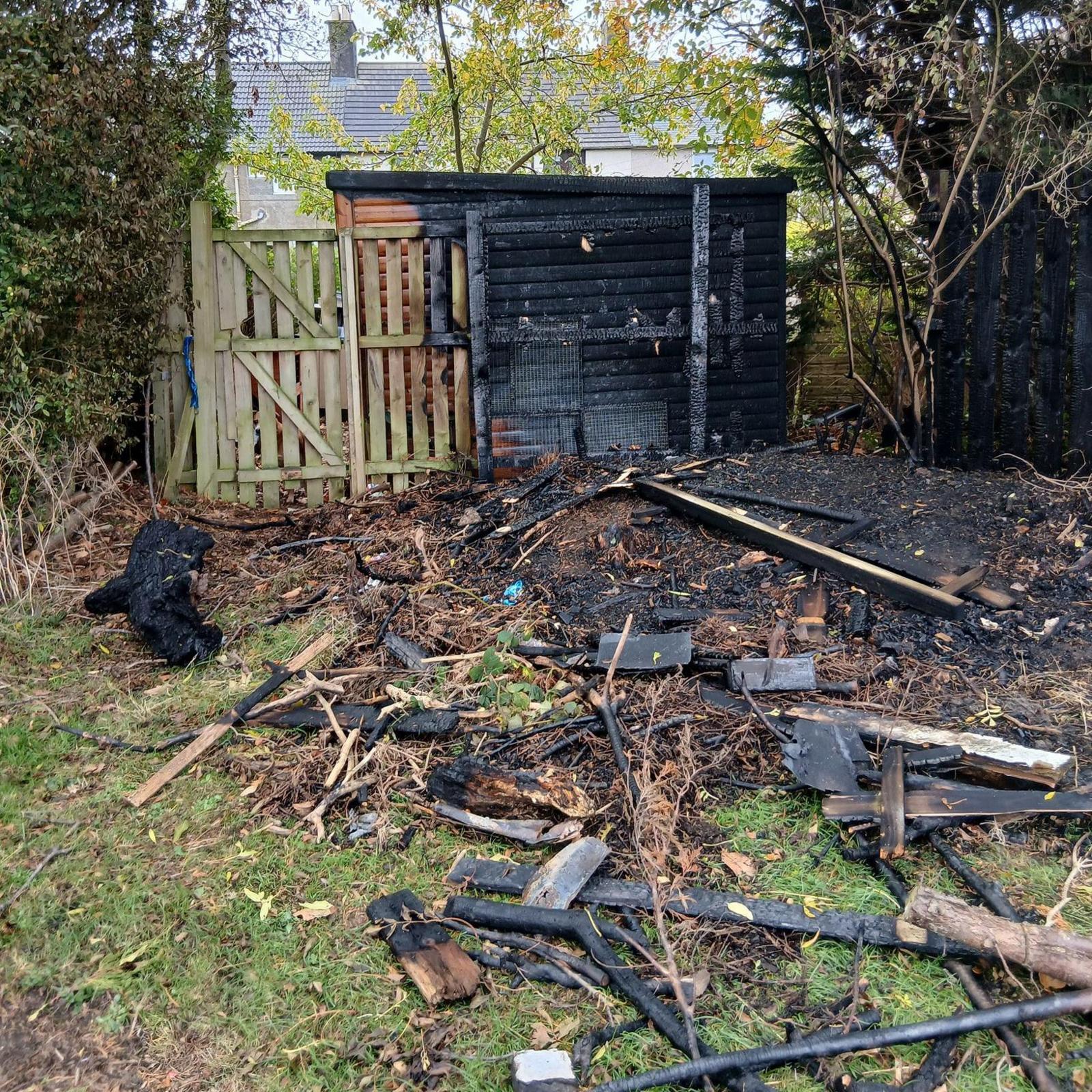 A burnt out shed with bits of burnt wood and rubble scattered on the grass in front of it