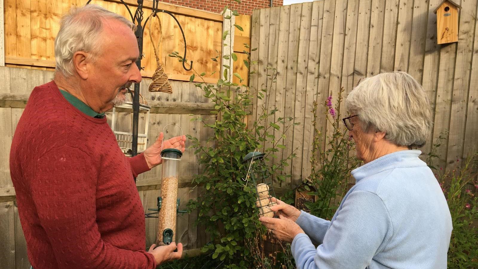 A man with short grey hair is wearing a red jumper. He is holding a bird feeder with seed in it. Behind him is a bird feeding hanger. Next to him is a woman with short grey hair, she is wearing a light blue top.