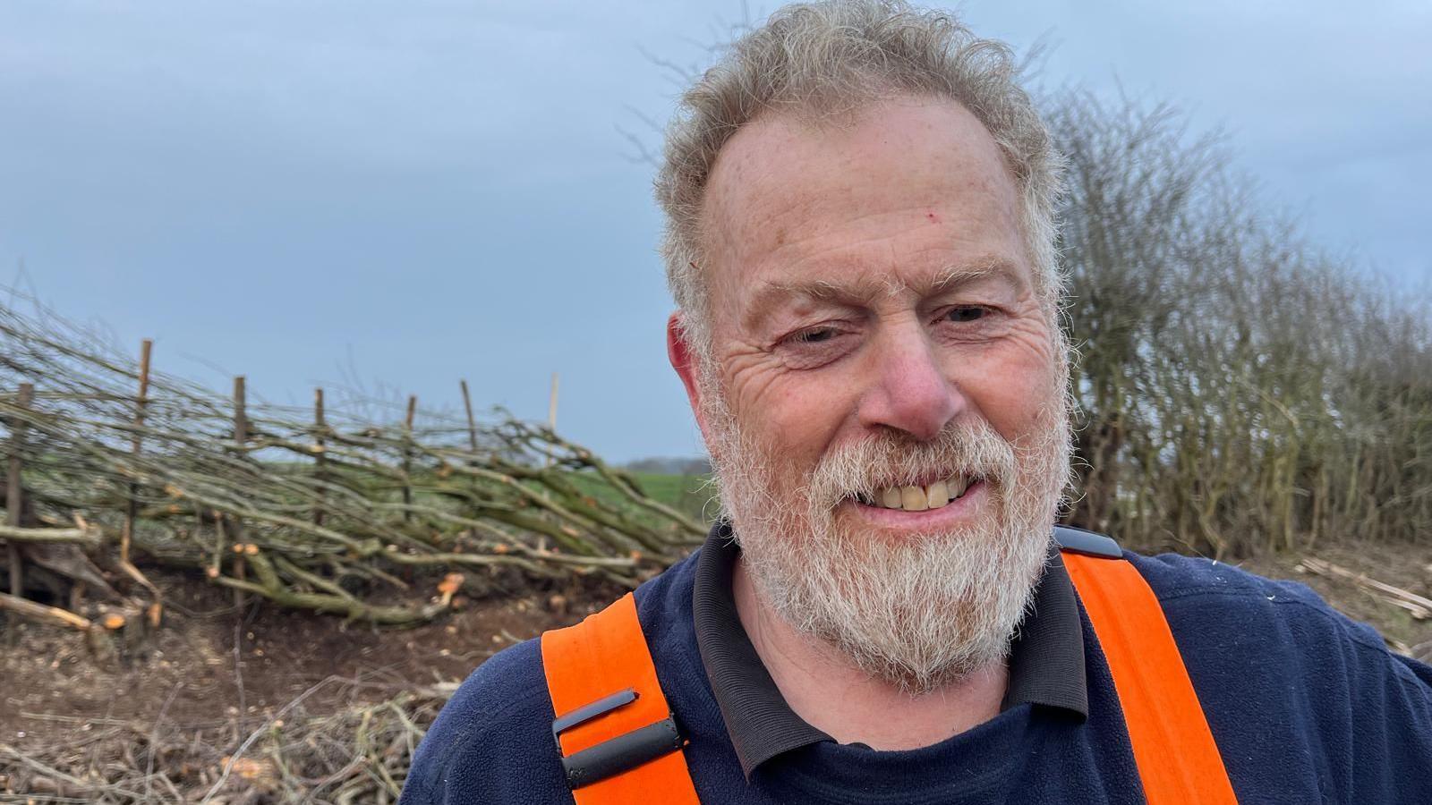 Steve smiles at the camera with a hedge chopped and laid down on one side and a hedge growing tall on the other side 