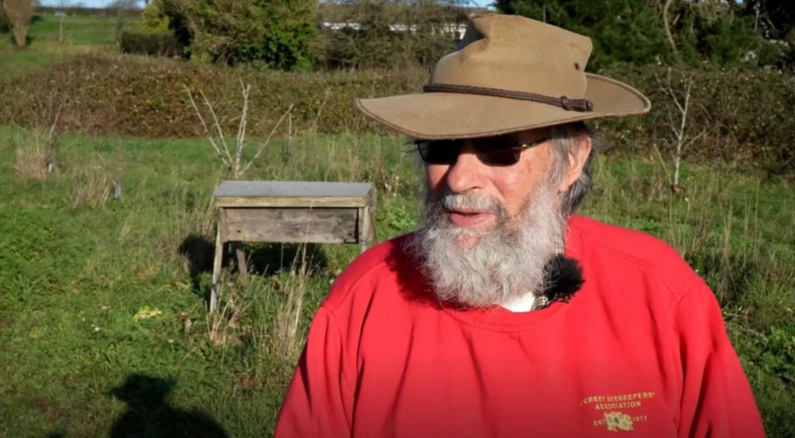 Bob Hogge is wearing a red jumper with the Jersey Beekeepers Association emblem on it. He had a white beard, black sunglasses and a brown wide brimmed hat. He is stood outside on a sunny day.