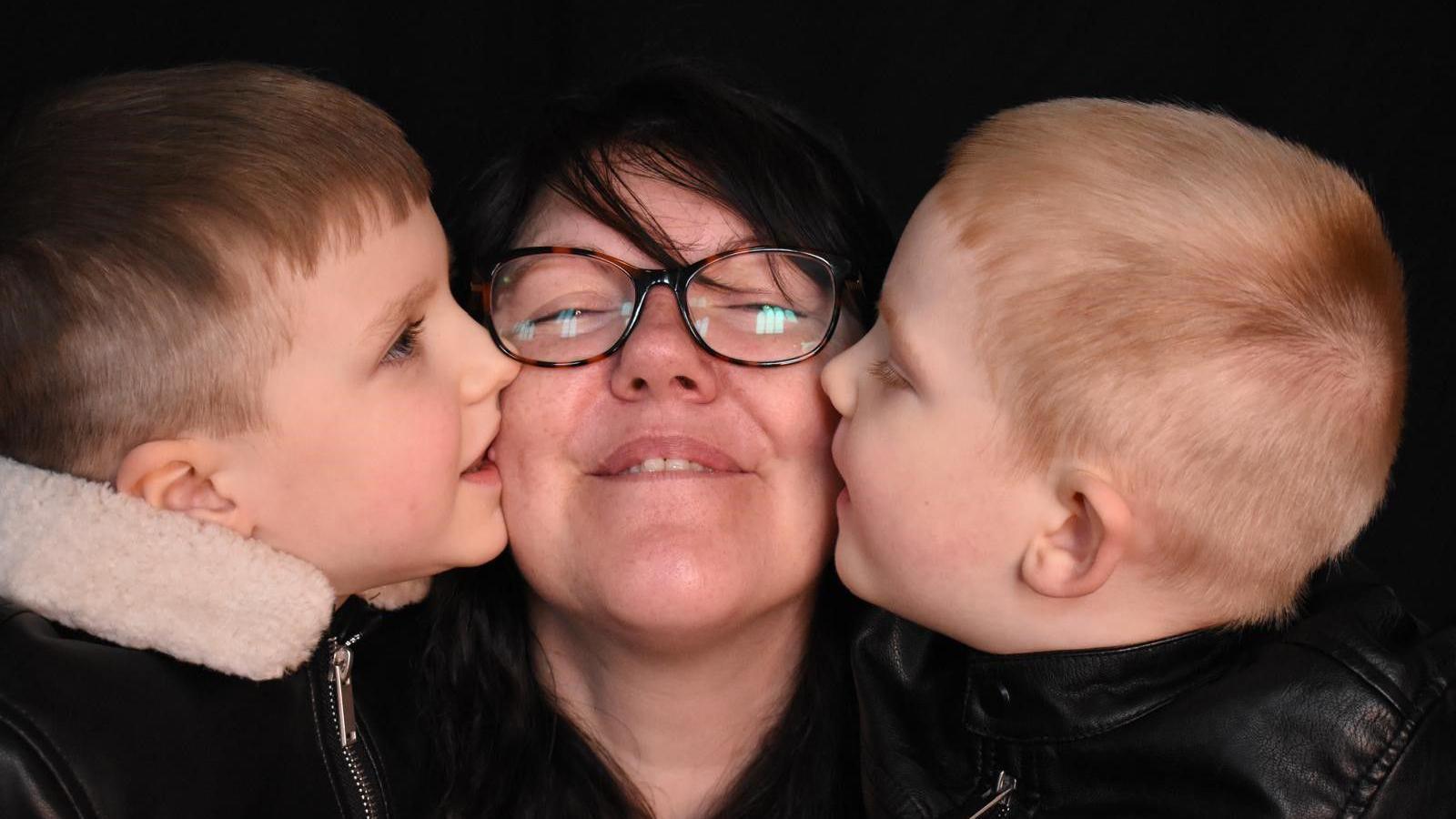 A dark-haired woman with glasses smiles at the camera while her two young sons, both with short blonde hair, kiss her one on each cheek.