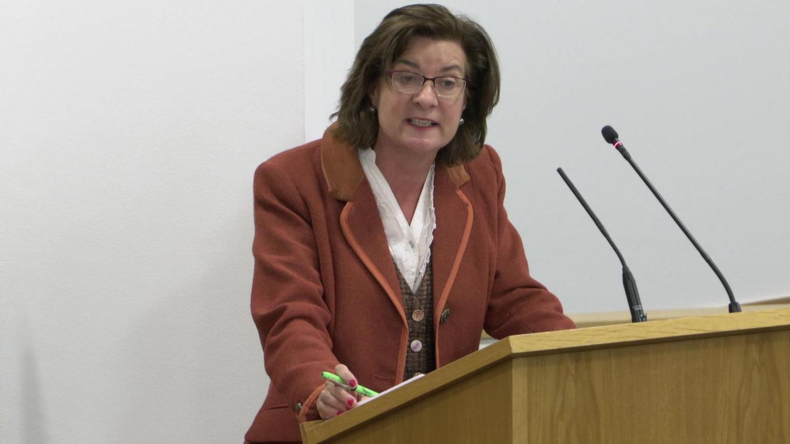 Eluned Morgan responding to Darren Millar during First Minister's Questions in the Senedd. She is standing at a light coloured wooden lectern with two microphones on it and holding a green pen in her right hand. 