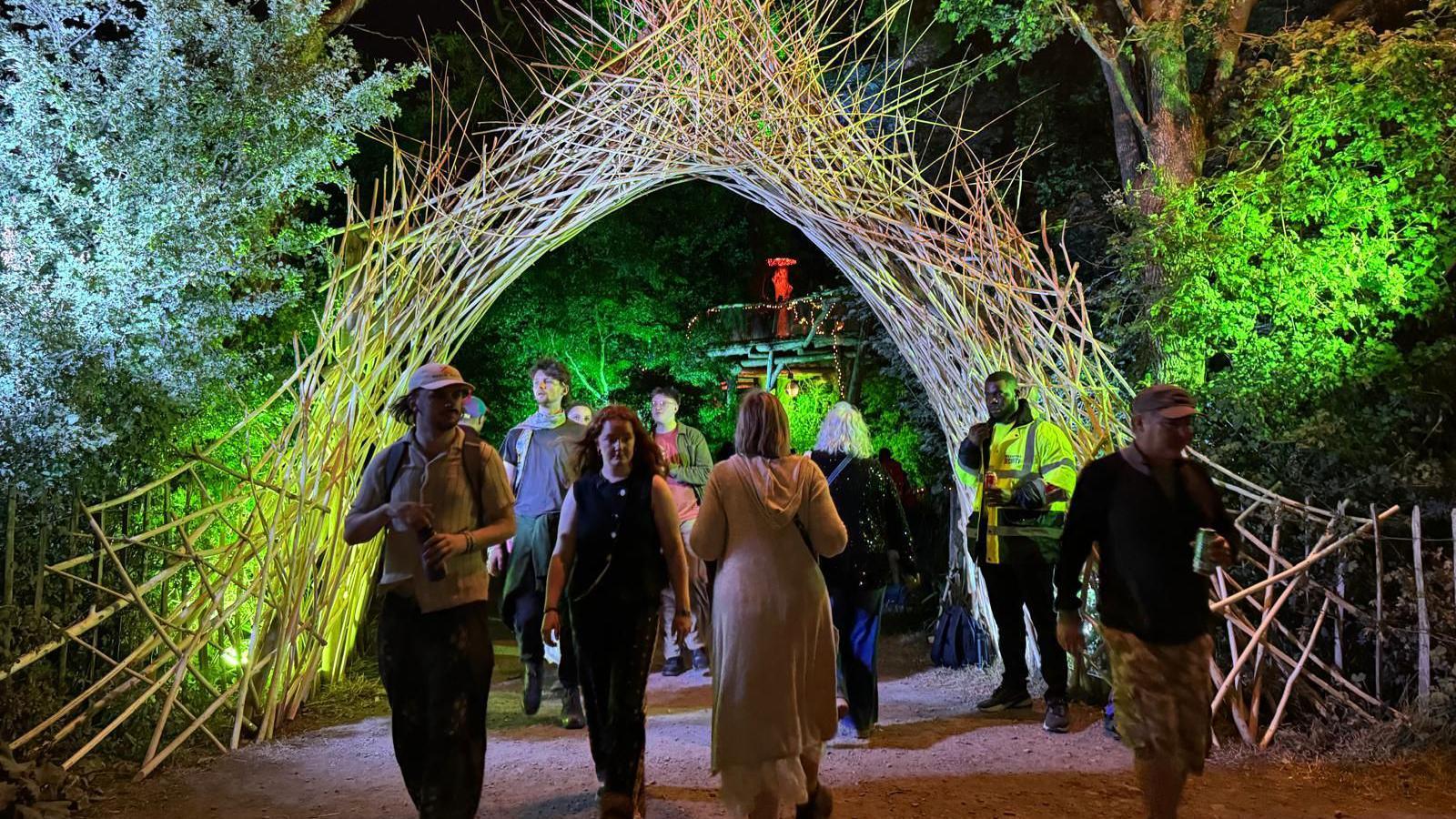 A wicker arch spans across a walkway in a wooded area. It is night-time and green lights are shining on the arch and trees. About a dozen people walk along the path.
