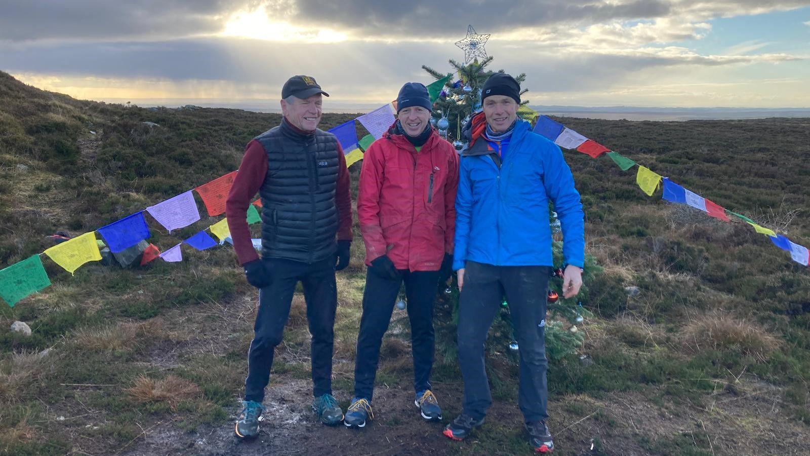 Three men stand in front of a Christmas tree which is secured by ropes covered in flags. The tree is on a remote hillside and the men are wearing mountain gear 