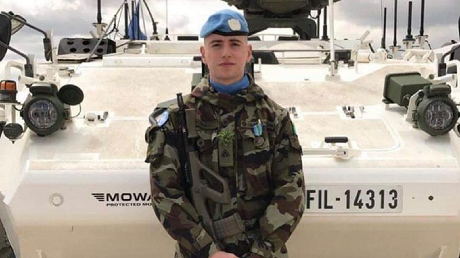 Irish soldier Seán Rooney, wearing camouflage clothes and a blue beret. He is holding a gun and standing in front of an armoured vehicle.