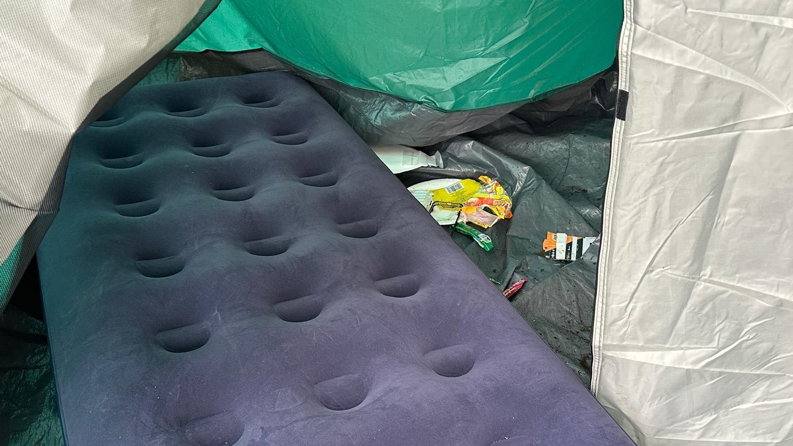 A camping mattress has been dumped in a tent. There is a blow-up camping mattress among other bits of litter left. 