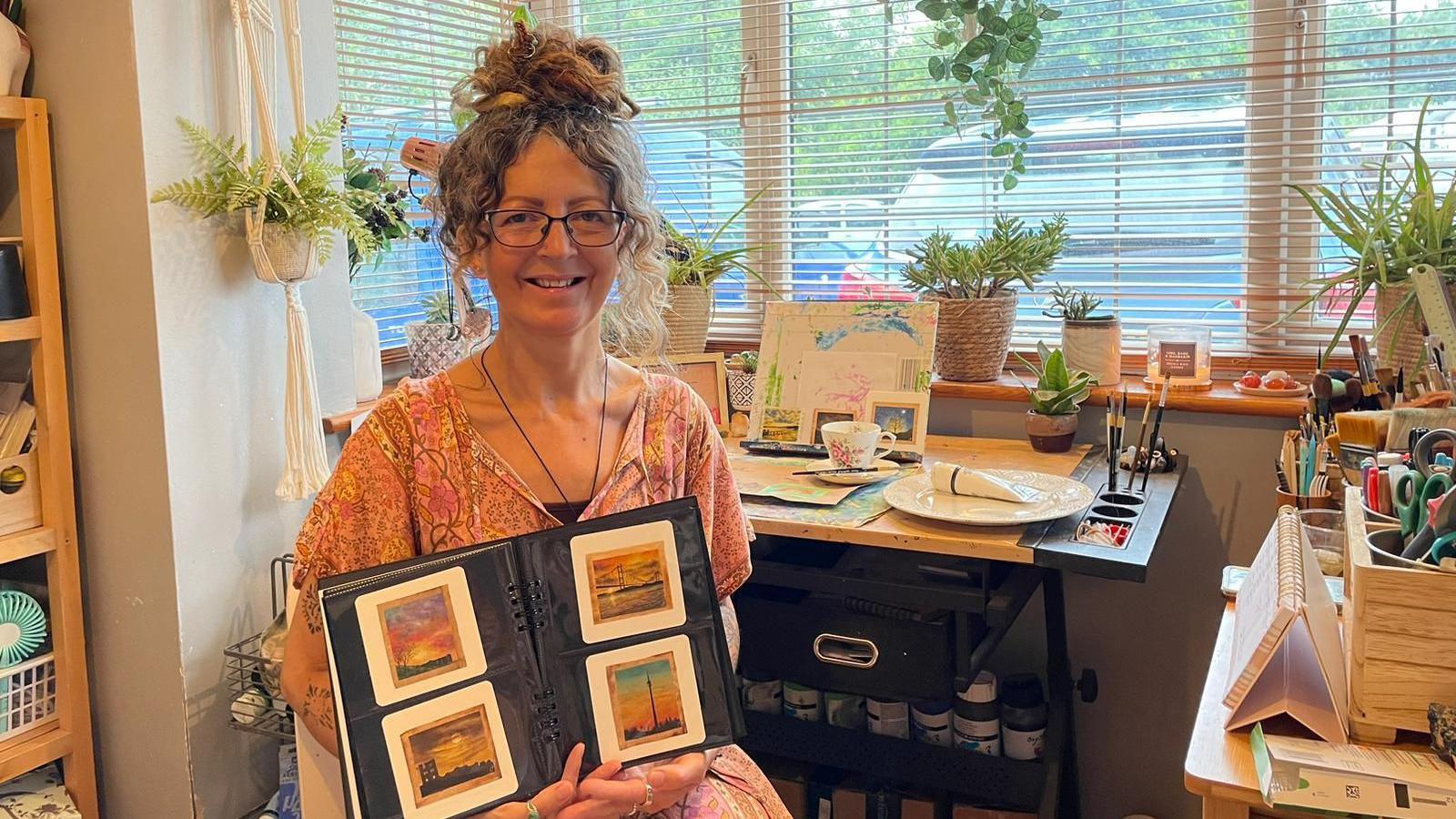 A woman wearing a pink patterned dress and glasses, sitting in an artist's studio and holding up a book with four tiny paintings in it, painted onto teabags. There's a desk and a window behind her, with plants on the window sill.