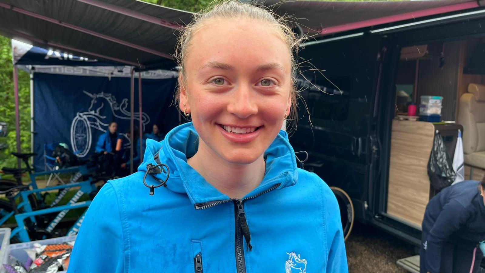 A woman in her early twenties wearing a blue tracksuit top. She is stood next to what appears to be a campervan with its door ajar. She is smiling into the camera.