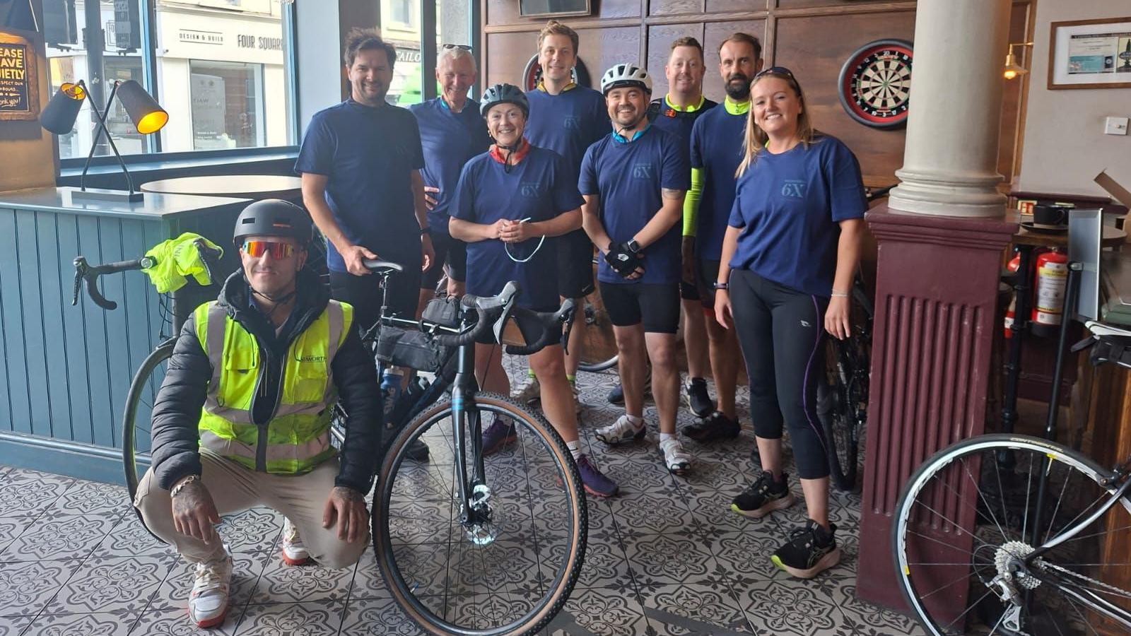 A group of people in cycling kit inside a pub, with a couple of bikes there as well. All are smiling at the camera, a few with helmets on too