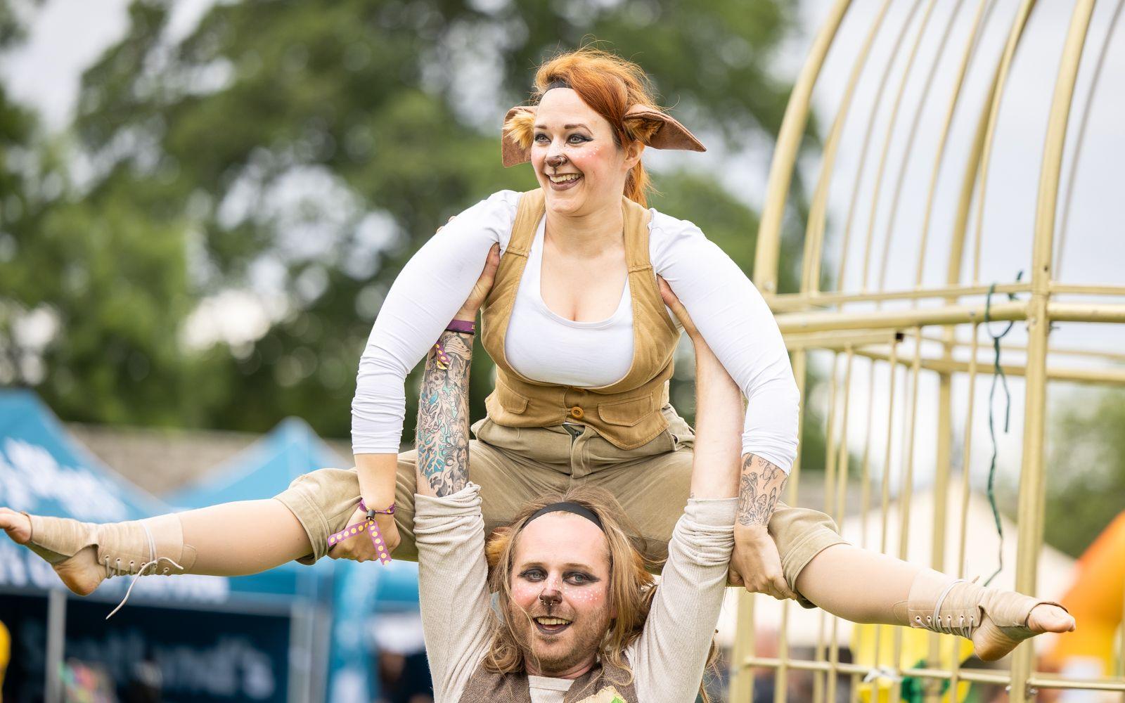 Costumed street performers wearing ears and face paint performing during the day, a man holds a woman doing the splits above his shoulders