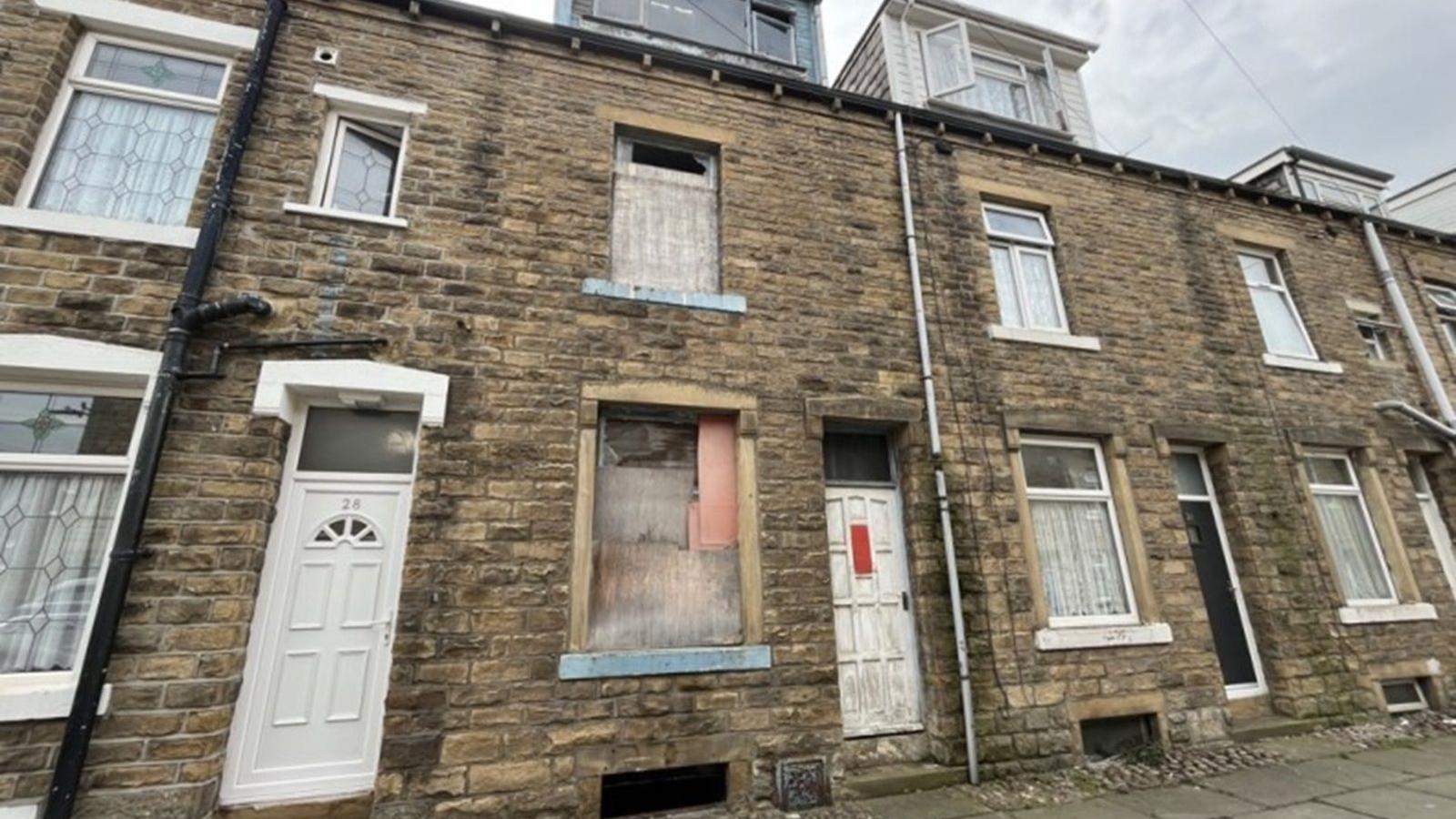 Row of stone-brick terraced houses on an overcast day. The central house appears abandoned or under renovation, with boarded-up windows and a worn white-painted door.