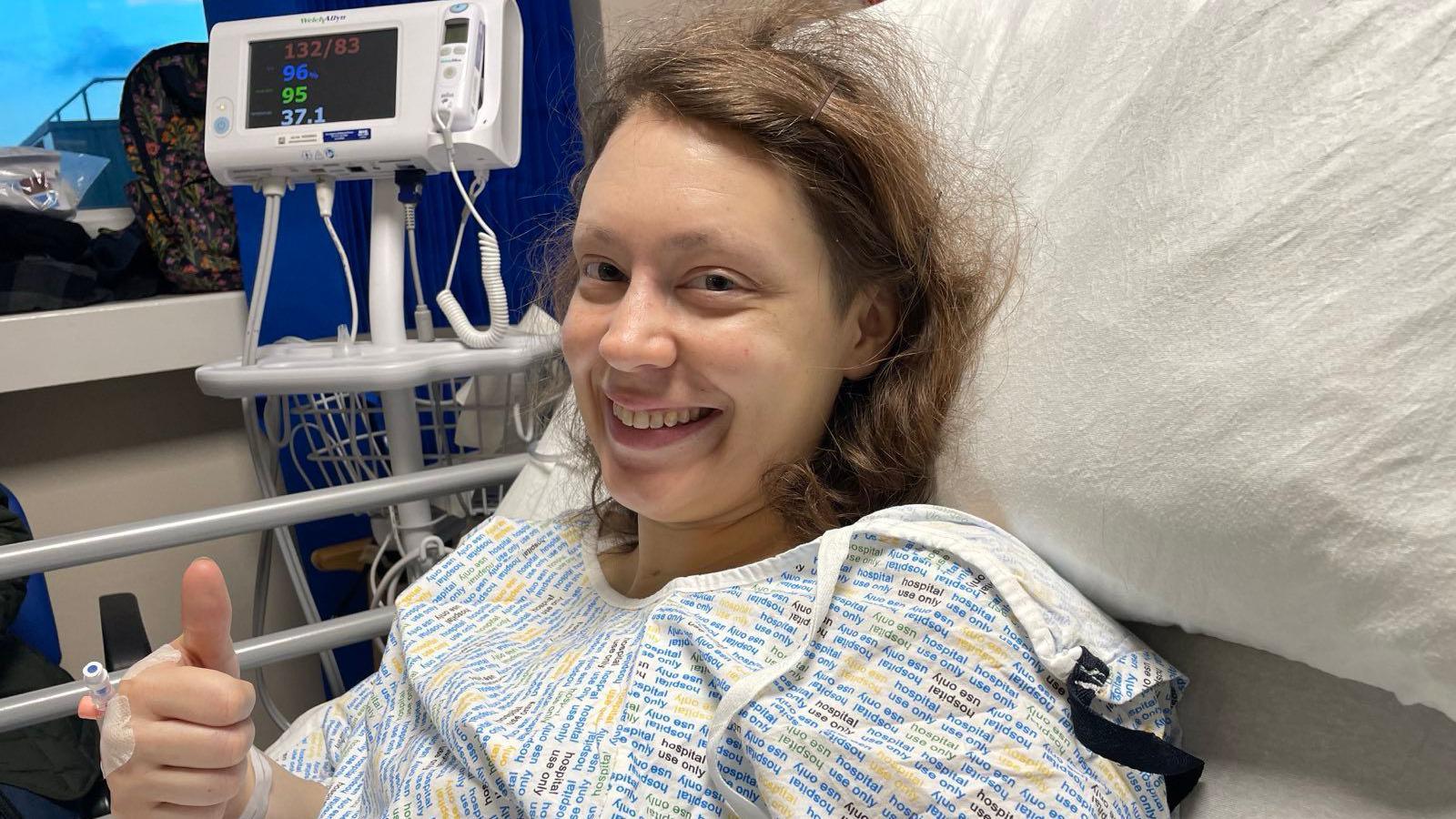 Kat is in a hospital gown in bed. She has brown hair and is smiling. There is a hospital monitor in the background.