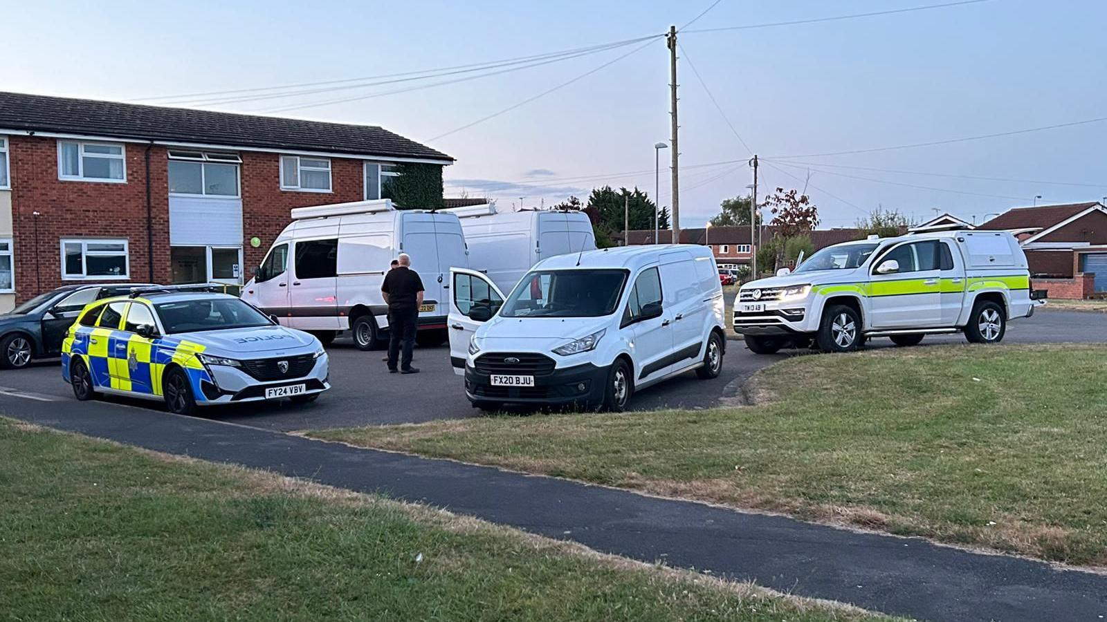 Police and military vehicles and two white vans parked with houses on the left