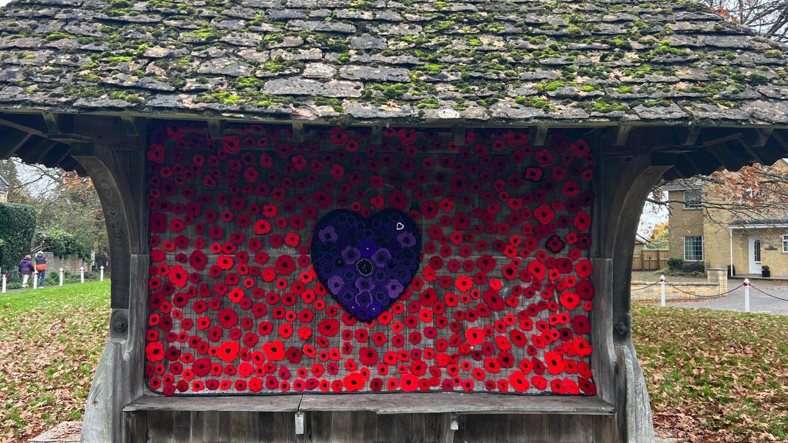 A bench with wooden shelter and back, a sea of red poppies with a purple heart with purple poppies in the middle