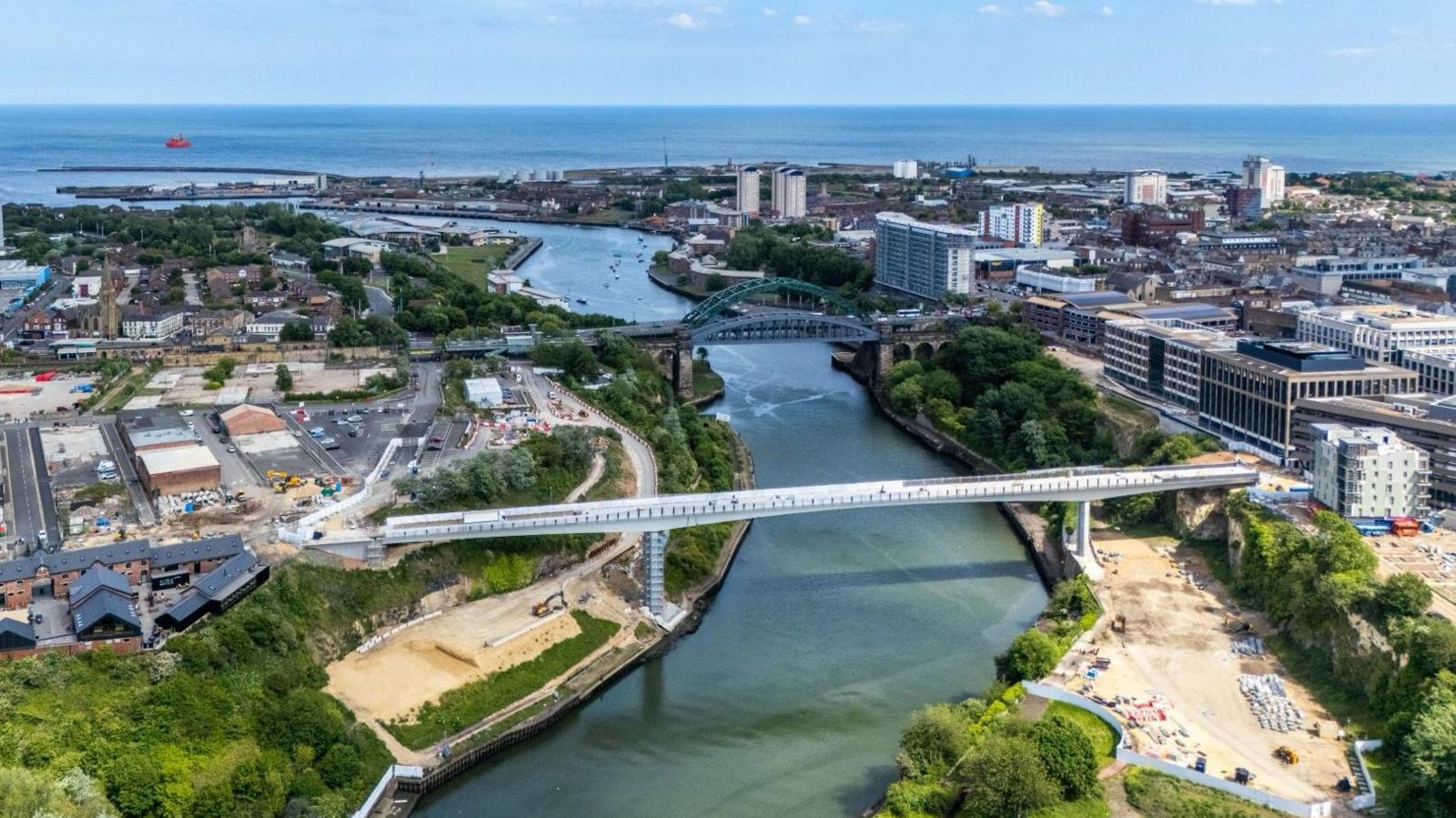 An aerial view of the footbridge. It is a long white structure crossing the River Wear and connecting Keel Square on the right to the Stadium of Light, with cranes next to it on both shores. Wearmouth Bridge and the North Sea are also visible in the distance.