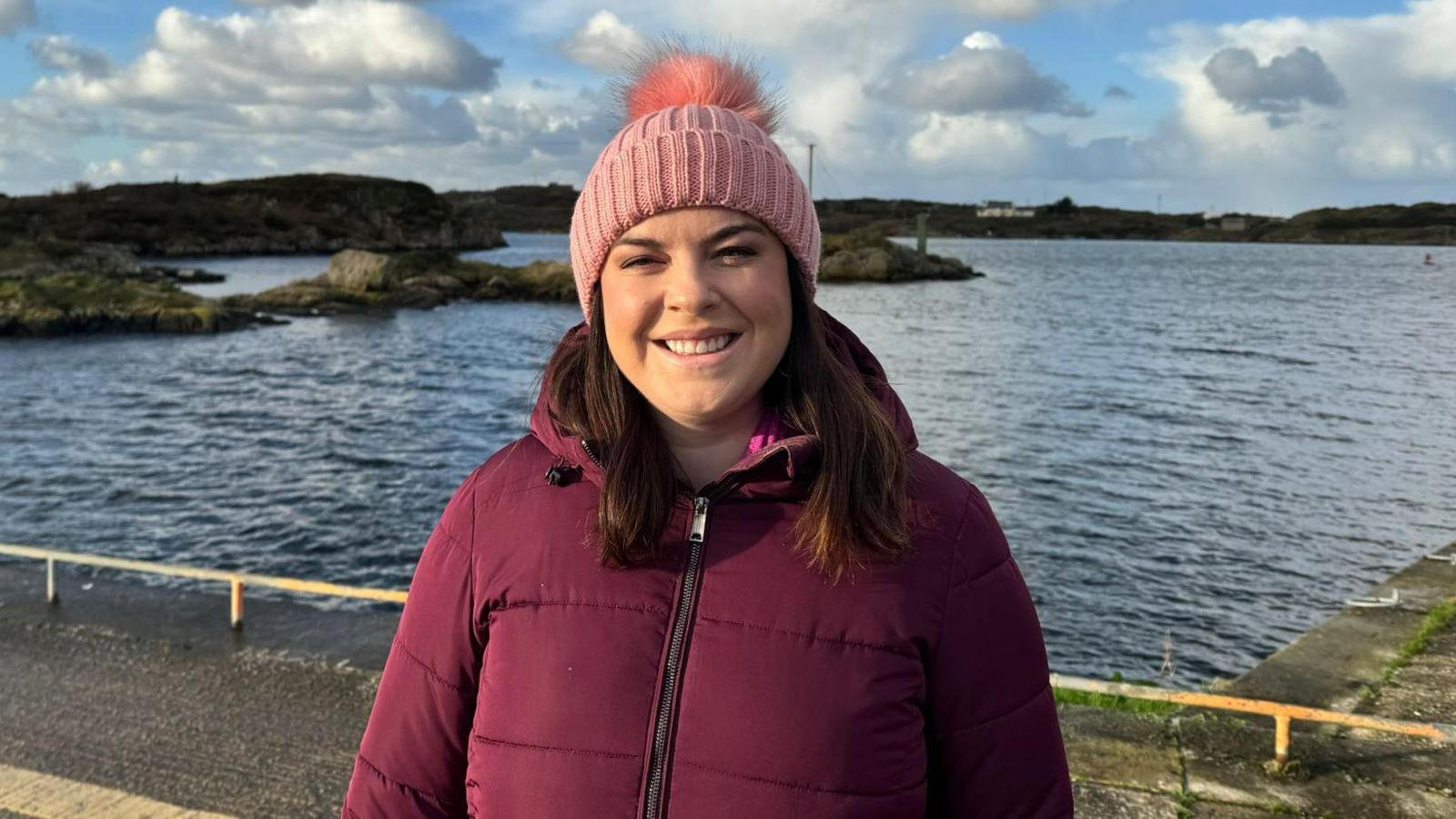 Áine Ní Bhreisleáin - a woman wearing a maroon coloured puffer jacket and a pink knitted hat with a pompom is smiling at the camera. She is standing on a harbour pier, with a body of water behind her.