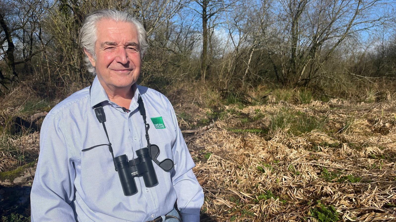 Tony Juniper poses for a photograph in a blue shirt with a green badge that says 'natural England' - he's wearing black binoculars around his neck and is standing in front of a grassy riverbank fringed by trees.