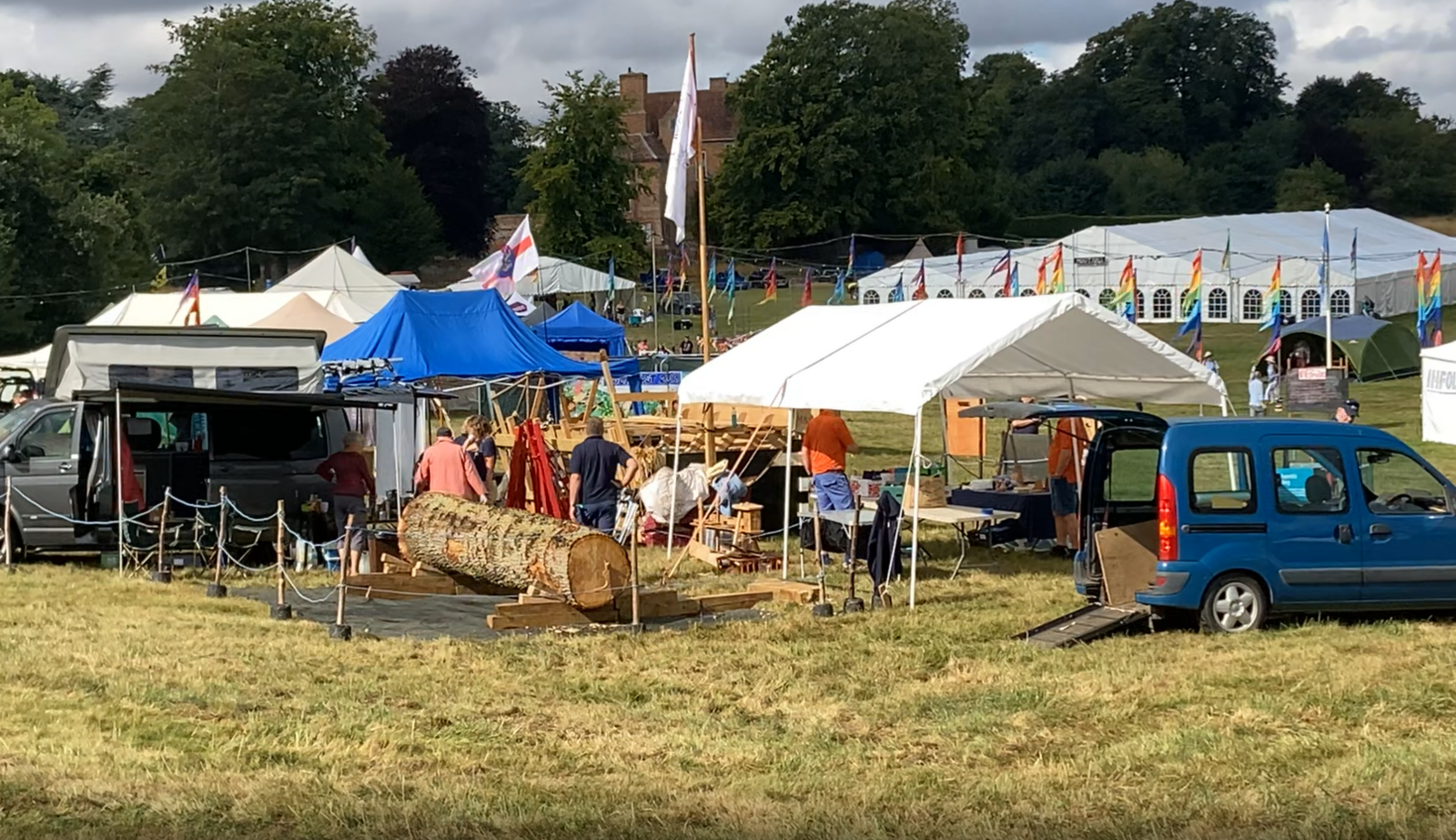 FolkEast attendees carrying items from parked cars into tents for the festival. There is a line of multi-coloured flags behind the tents. 