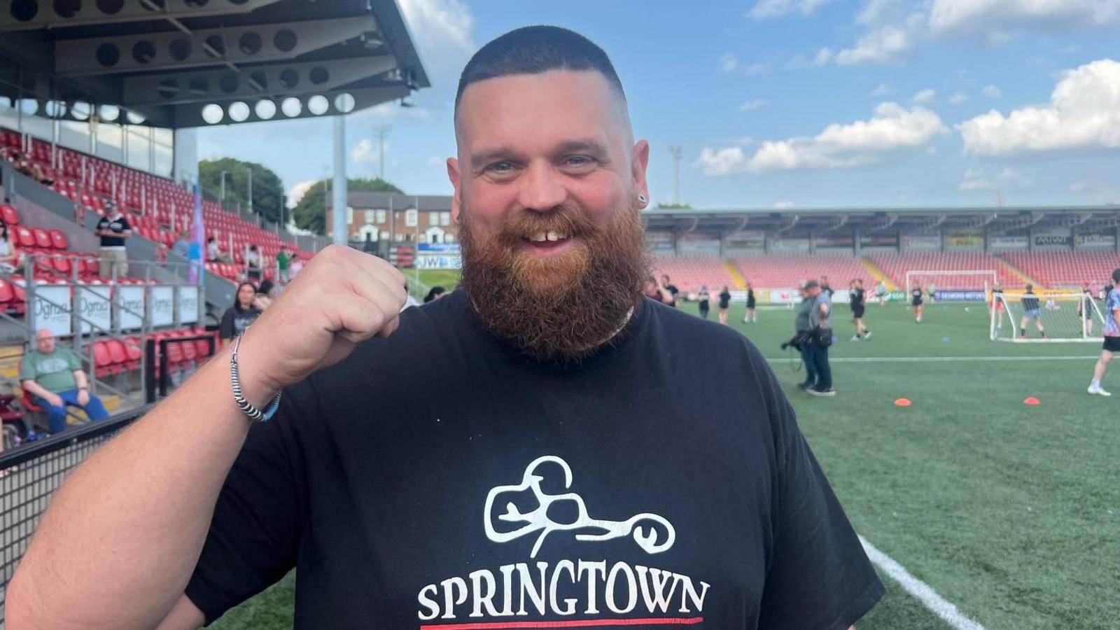 Daran McCann with a full reddish beard raises a clenched fist. He is standing in front of the stand in a football ground, wearing a black T-shirt. He has short, tightly cropped dark hair.