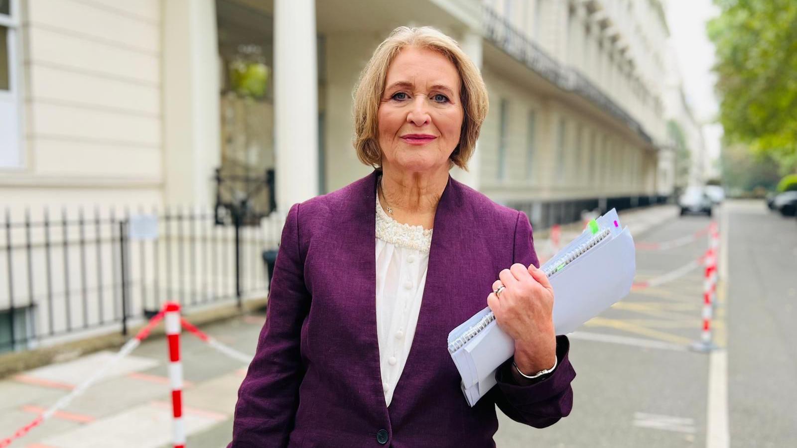 Baroness Anne Longfield stands, with papers in her hand, wearing a purple blazer and white blouse. She is standing outside a white brick building.  