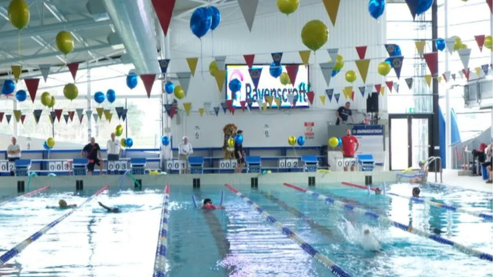 A picture of a number of lanes in a swimming pool. There are lots of brightly coloured bunting and balloons above the pool. There are swimmers in the pool and there is a big TV screen in the background. 