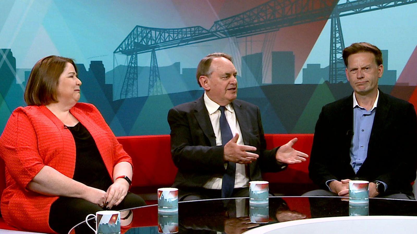 Julie Minns, Lord Kirkhope, and Andrew Husband sitting on the BBC Politics North red sofa. Minns, with a brown bob cut and wearing an orangey red half sleeve jacket and black top looks to her left with her face contorted slightly as if she is mid laugh or cry. In the middle with receding but still mainly light brown hair, Kirkhope wears the standard dark suit, dark tie, light shirt combo. He's mid sentence and has both hands stretched out in front of him. His expression could be that of someone hoping to convince, but might be exasperation. Husband has gone office casual. Black jacket over a blue shirt with the top buttons undone. Grey trousered, no tie, neat brown hair that has a fair amount of volume. With his hands lightly clasped in front of him, his mouth is turned up at one side, perhaps showing a lack of agreement with Kirkhope, or perhaps just confusion. There are four BBC Politics branded mugs on the table in front of them, perhaps for thirst avoidance, perhaps for aesthetically repetitive marketing.
