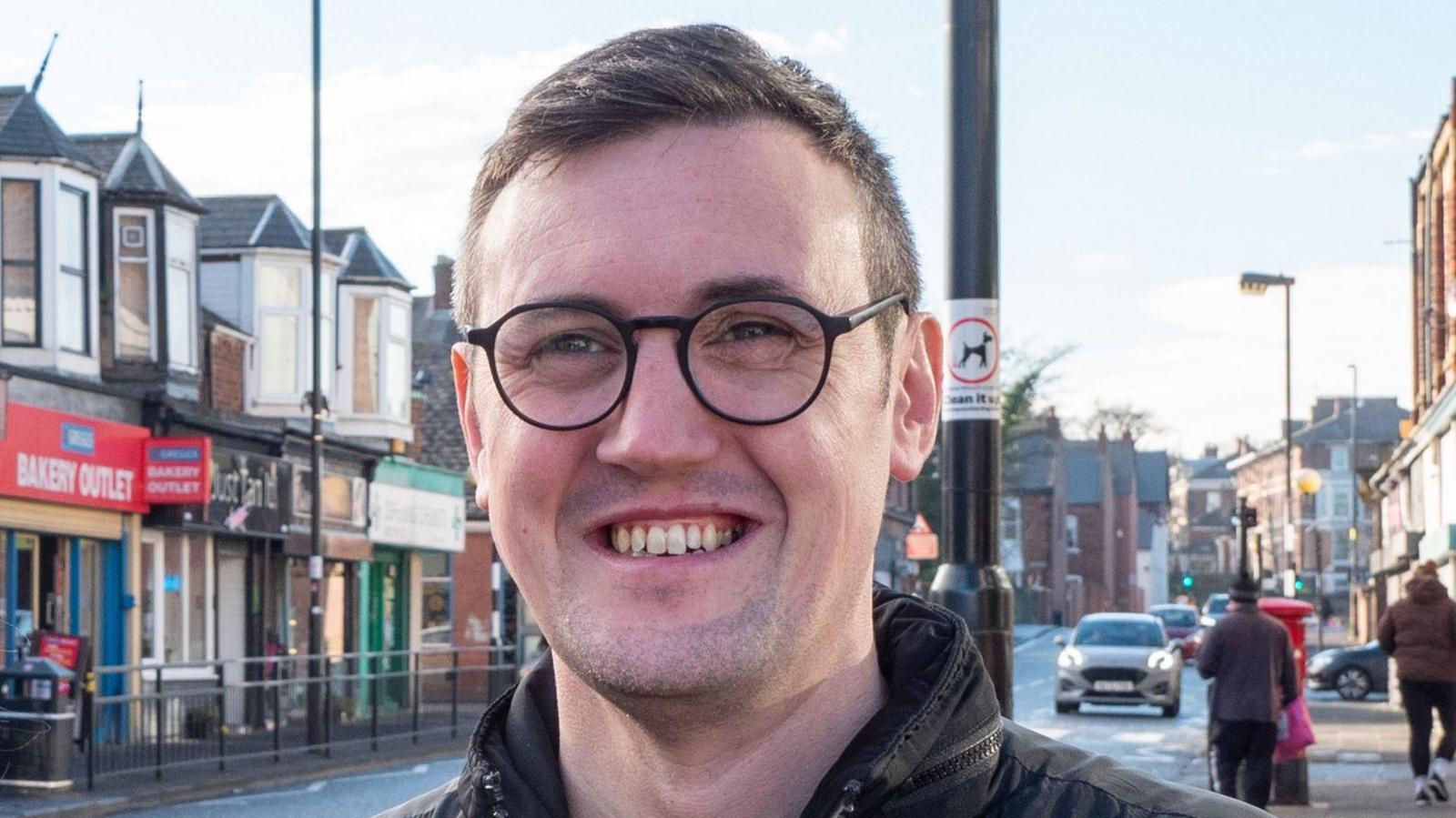 Councillor Michael Mordey is stood on a high street and is smiling at the camera. He has short brown hair and round glasses.