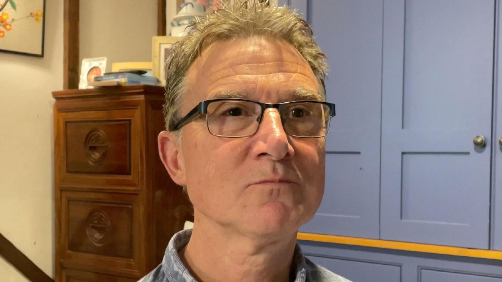 Mr Reid looking to the right of the camera, unsmiling. He is photographed inside a property with purple cabinetry behind him and a dark wooden chest of drawers. He has glasses and short grey hair. 