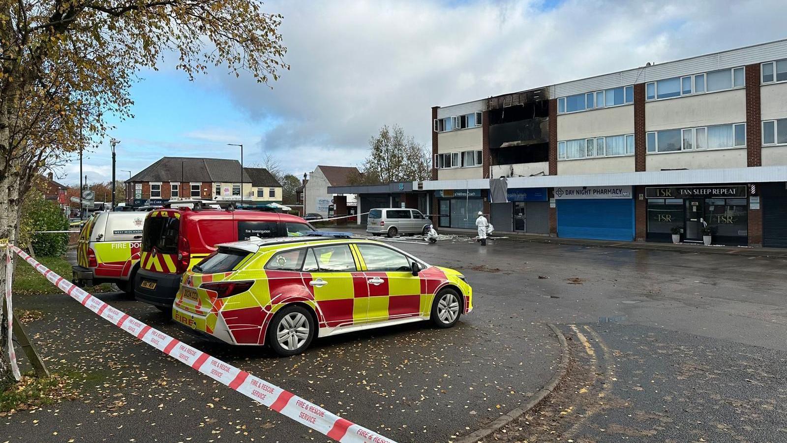 Three fire service vehicles, parked next to each other, are set behind a red and white tape. In the background is a building which has extensive burn marks from an explosion which has taken place in the flats above a row of shops.