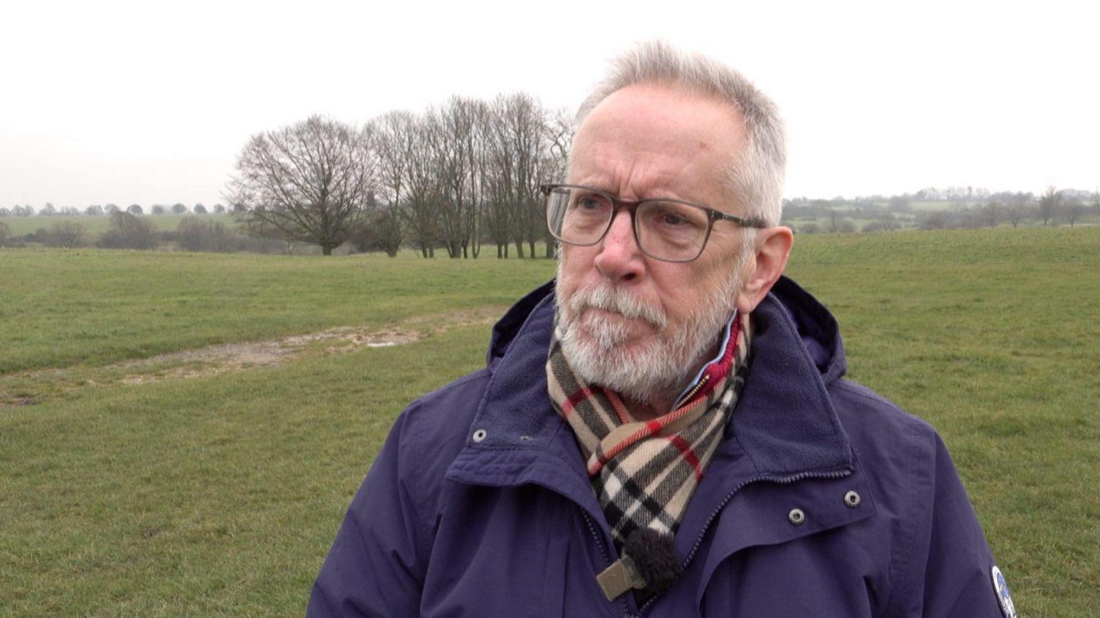 A man with short grey hair and a short grey moustache and beard wearing glasses and looking seriously towards someone next to the camera. He is wearing a purple coat and tartan scarf, with rolling fields behind him.