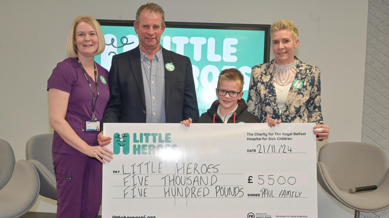 Cody with short blonde hair, square glasses and black hoodie holding up a cheque. He is standing alongside a man with short dark hair, blue shirt and black blazer, a lady with a blonde bob and purple scrubs, and a lady with short blonde hair, patterned suit and white top.
