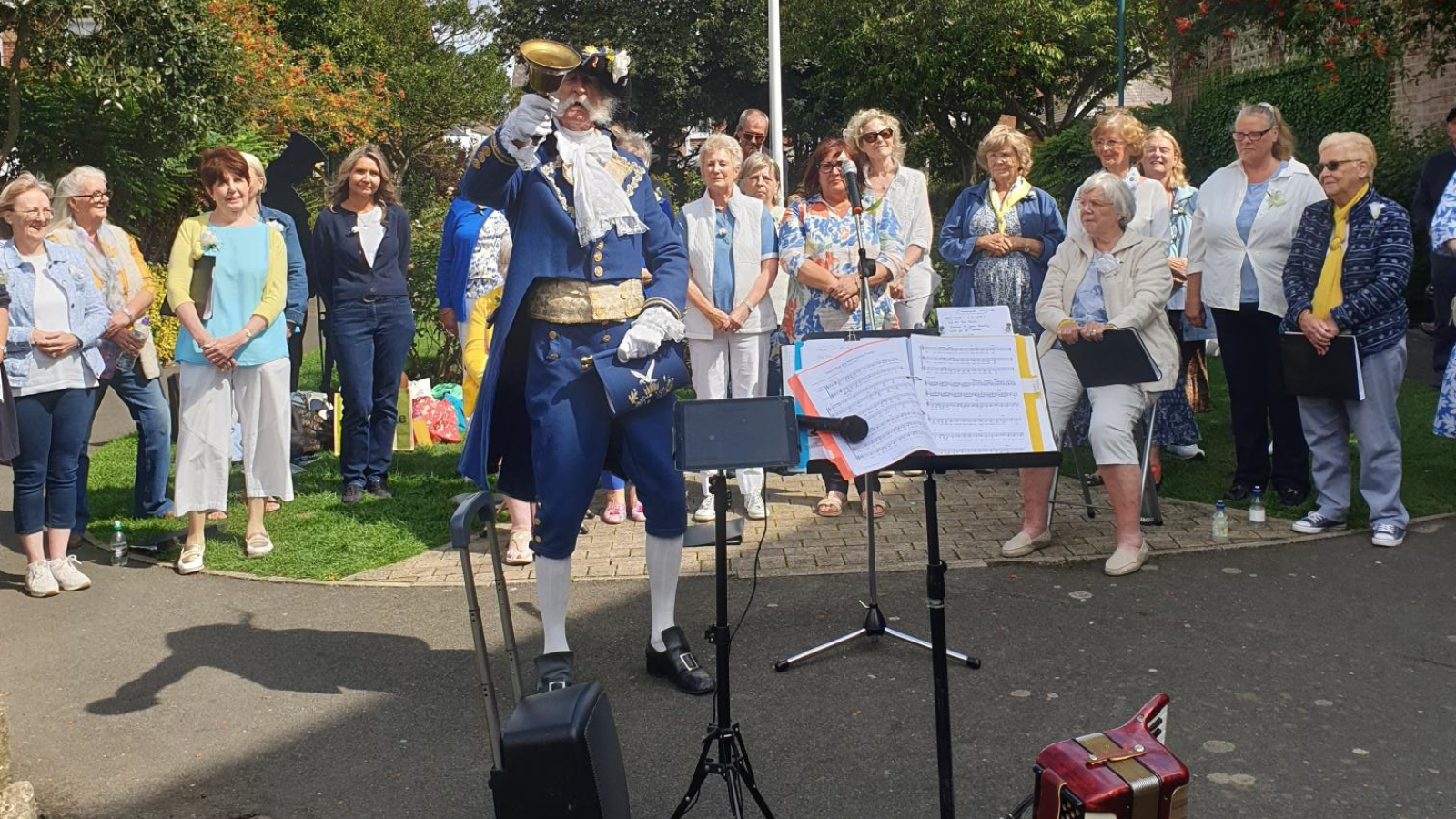 David Bull, in his town crier uniform, rings a bell to signal the start of a concert, surrounded by a mixed group of women. 