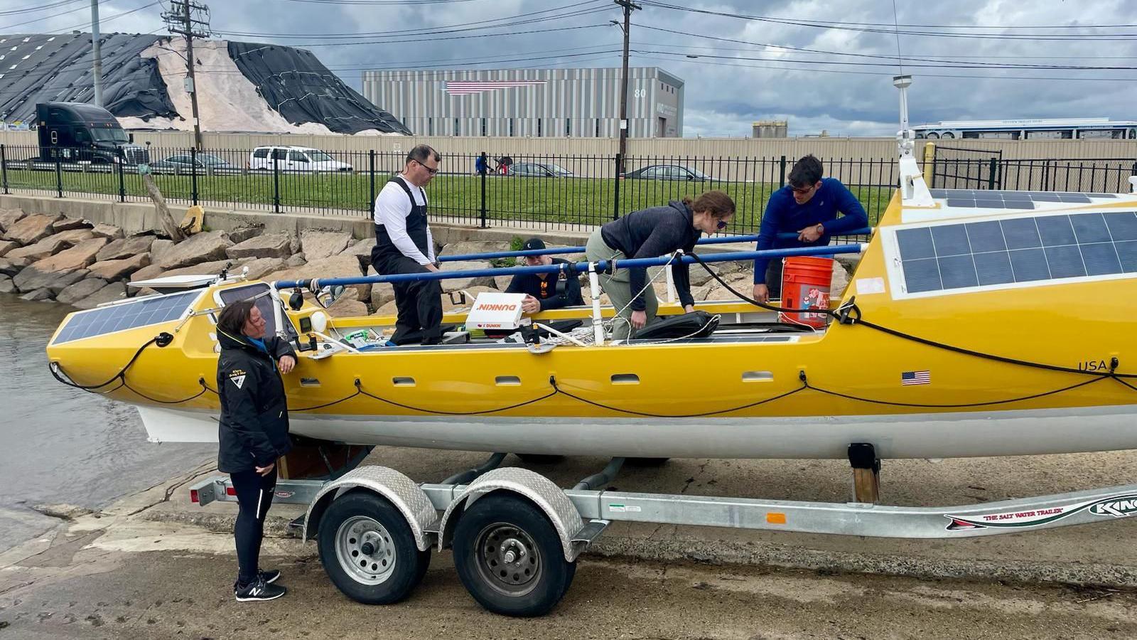 Three men and two women readying a yellow rowing boat, which has solar panels on the front, and stocking it with equipment. The boat is on a metal trailer next to the water.