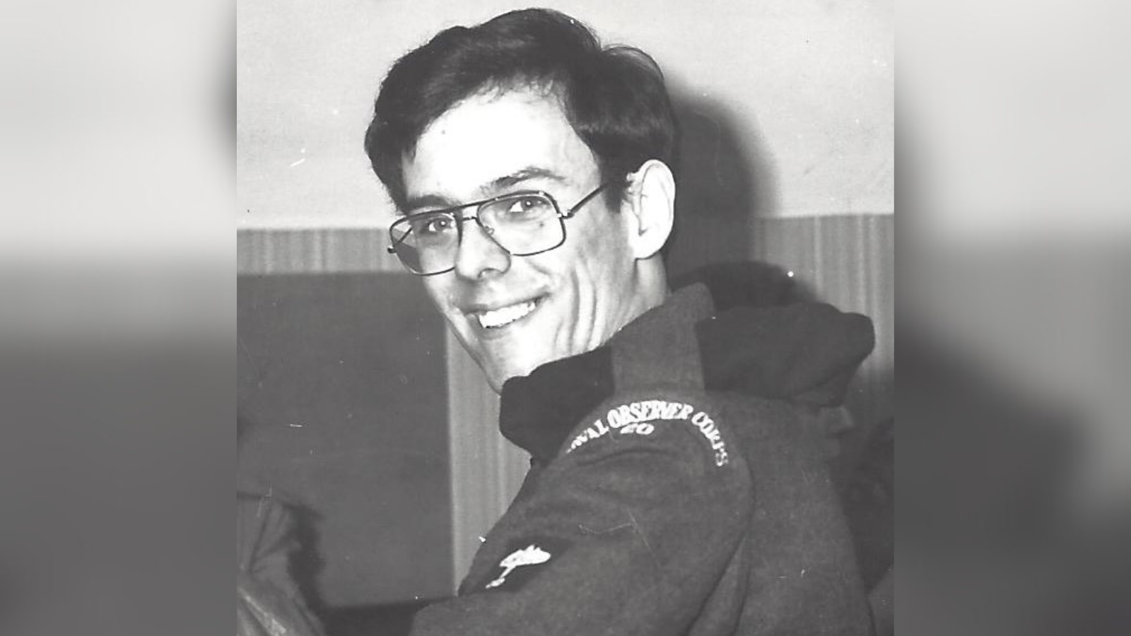 A black and white photograph of Tim Kitching as a young man, wearing his Royal Observer Corps uniform. He has dark hair and large, black framed glasses.