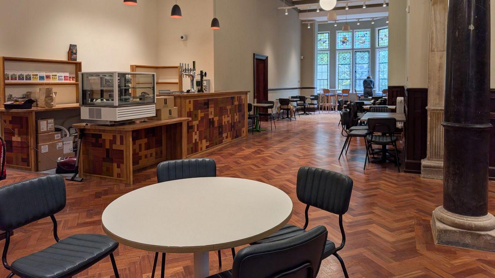 The café and bar inside the town hall. There is a bar to the left with beer taps and a unit to hold cakes. There are several empty tables and chairs. There are some stained glass windows at the far end of the room, where a person appears to be looking outside.