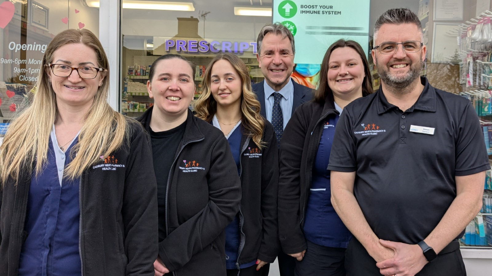 West of England Mayor Dan Norris, wearing a dark grey suit with blue shirt and dark blue tie, stands at the back of a group of people. The rest of the group are staff members at the Cadbury Heath Pharmacy and Health Clinic and are wearing branded black fleeces. They are a mixture of men and women