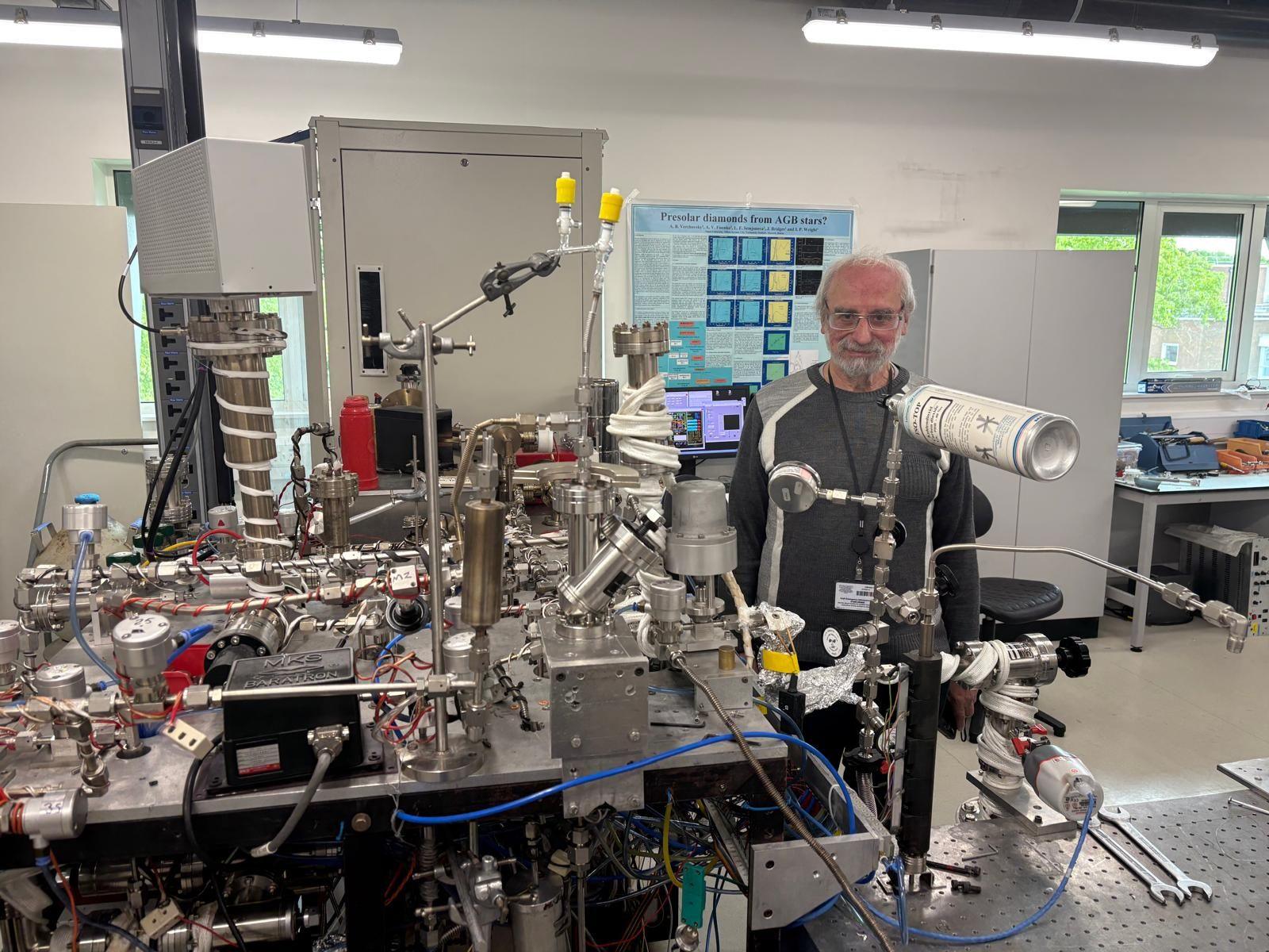 A large network of tubes, metal wires, plastic cables on a table in a lab. Sasha Verchovsky stands behind the machine, which is called Finesse.