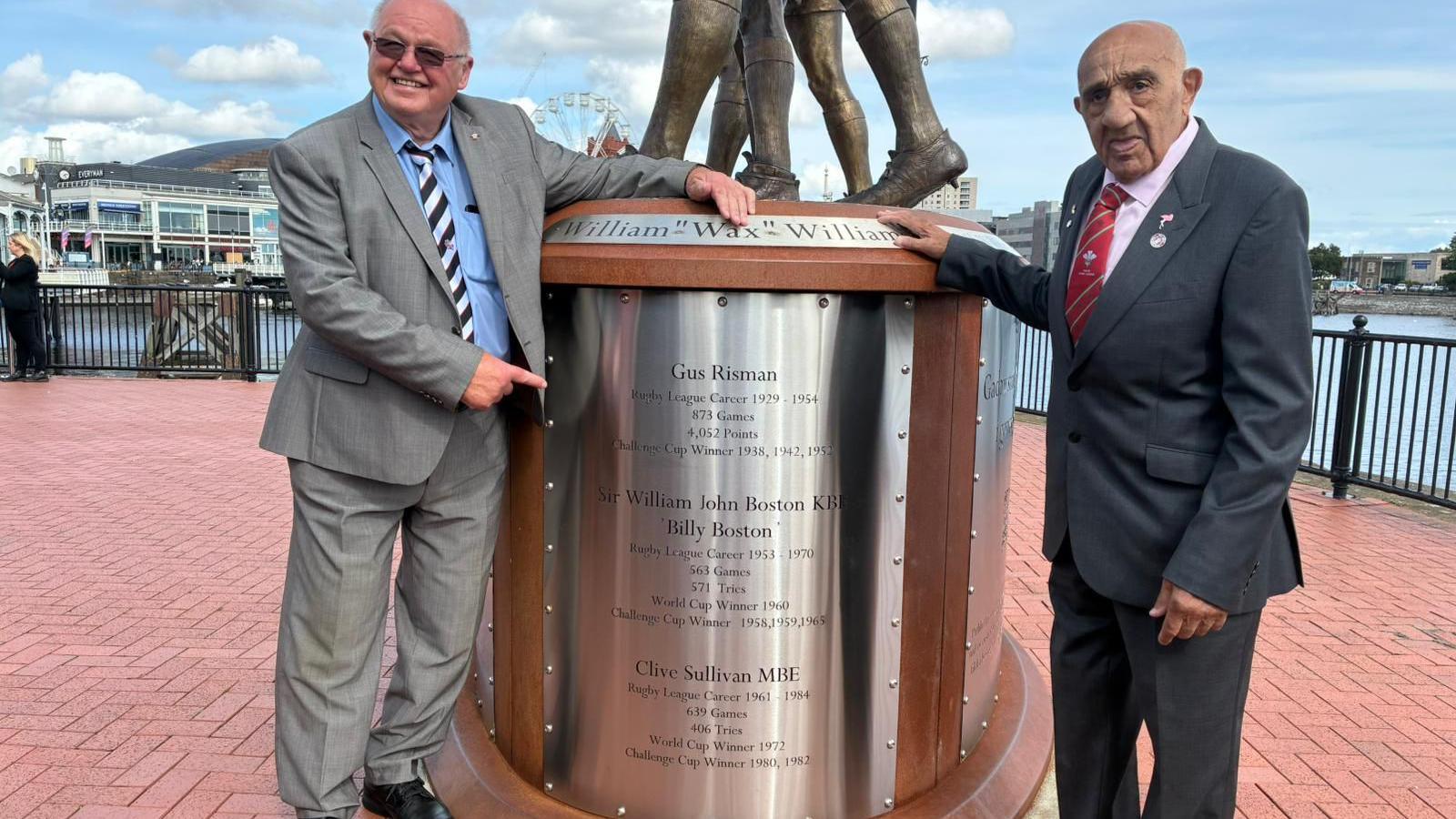 Two men stood either side of a statue smiling. Cardiff Bay can be seen on the left. There are three names engraved into the statue: Billy Boston, Clive Sullivan and Gus Risman. The man on the left is pointing and has a grey suit on, blue shirt and black and blue striped tie. He has shades on. The man on the right has a dark grey suit, light pink shirt and red striped tie.