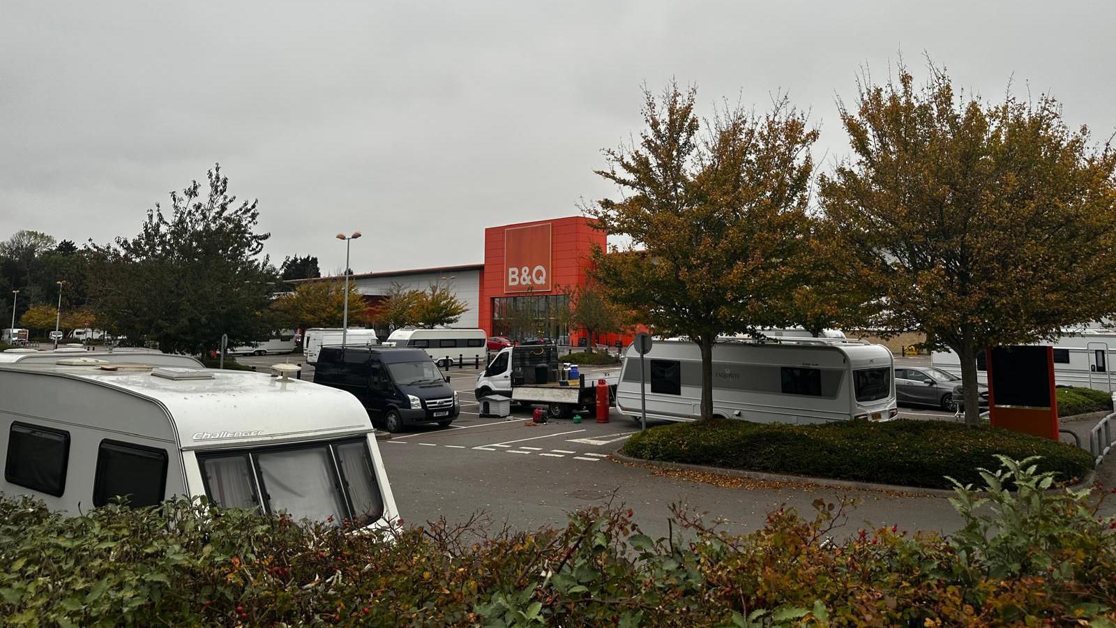Several caravans parked in a car park outside a B&Q store in Patchway, near Bristol. There are trees and hedges in the car park and the orange storefront can be seen in the distance.