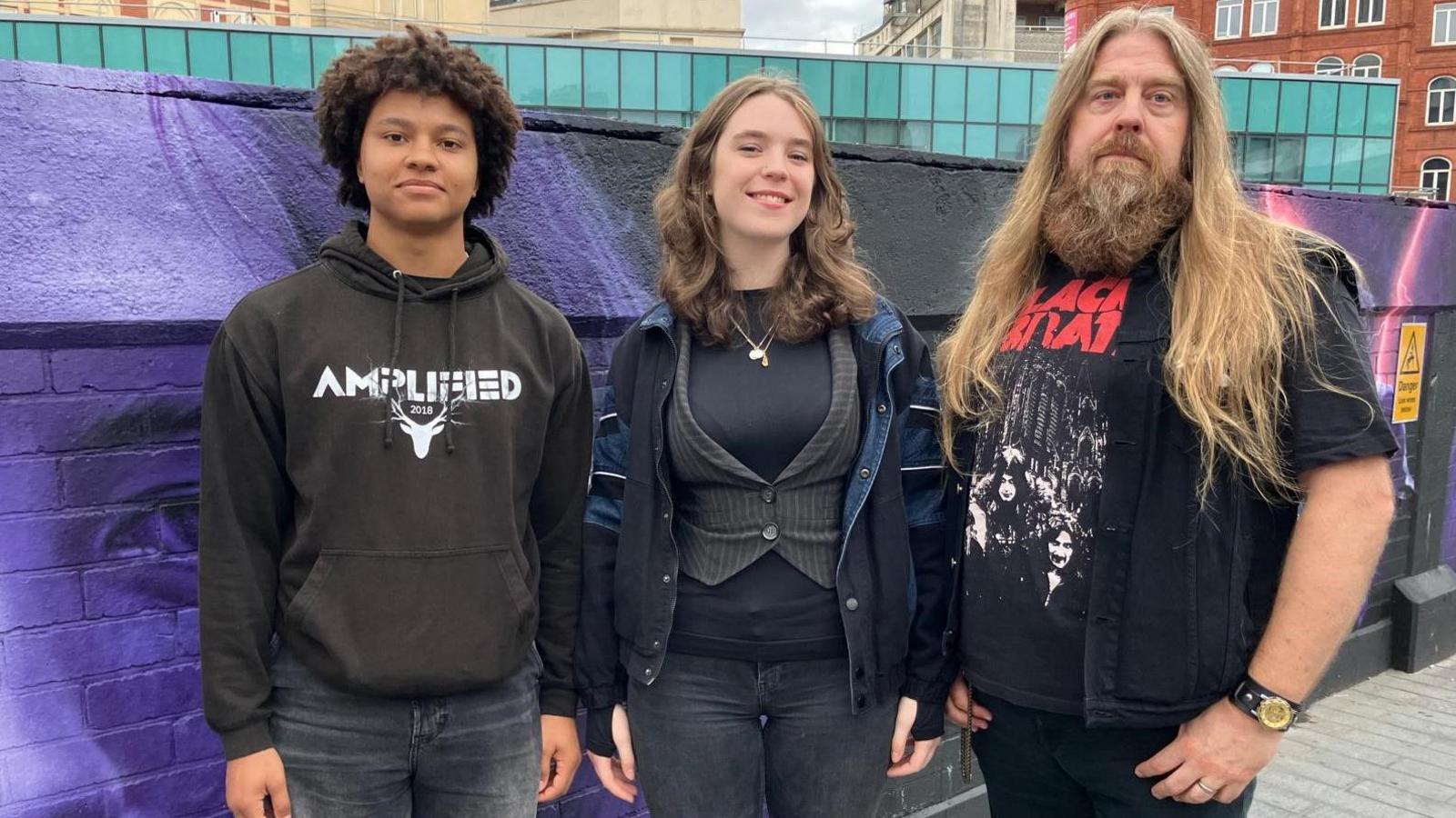 A man with long blond hair and a beard on the right, next to two others. Part of the purple, pink and black mural can be seen behind them on the wall.