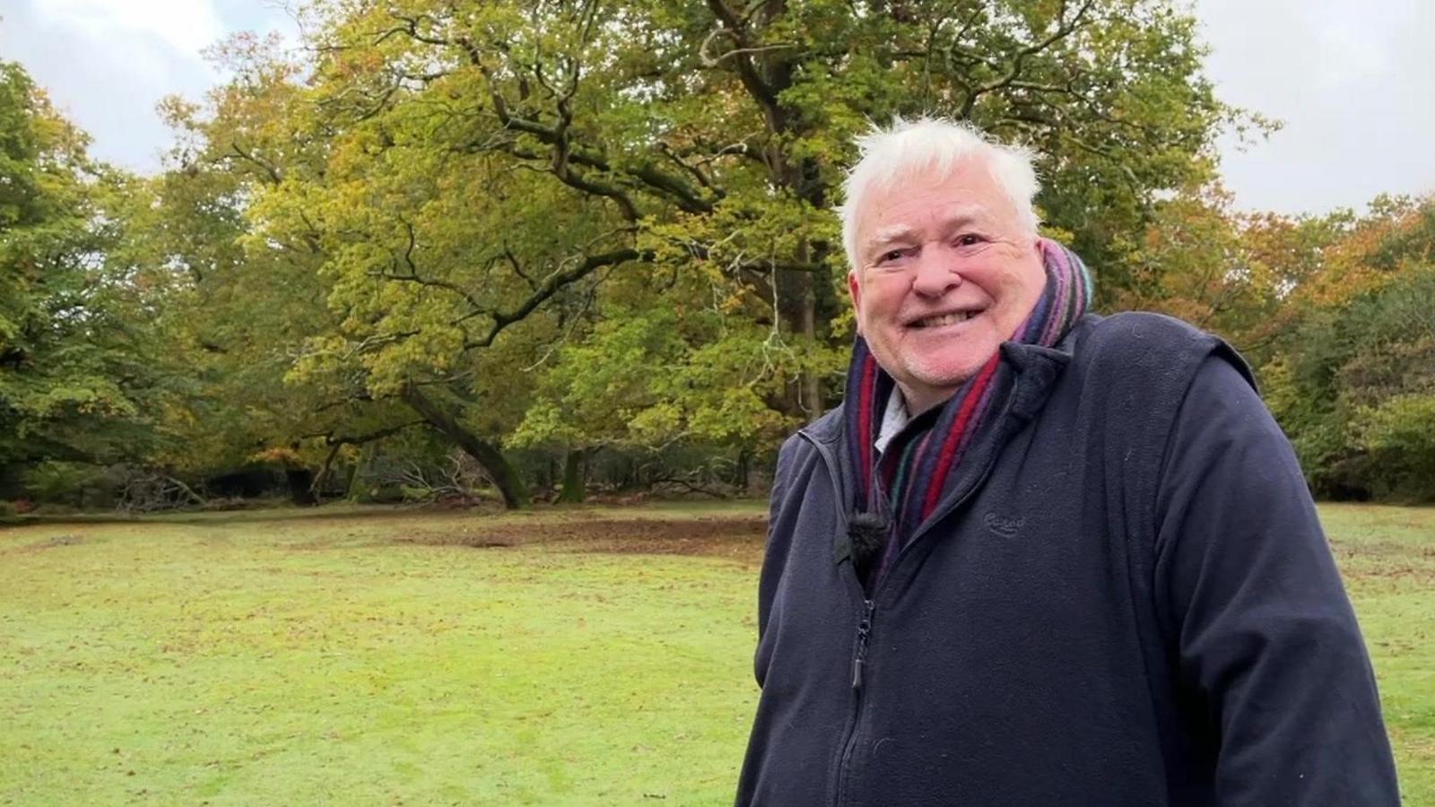 Bill Reynolds standing smiling wearing a dark blue fleece and striped scarf. Behind him is a wooded area of oak and beech trees with leaves which are starting to turn to yellow and brown but are still mainly green.