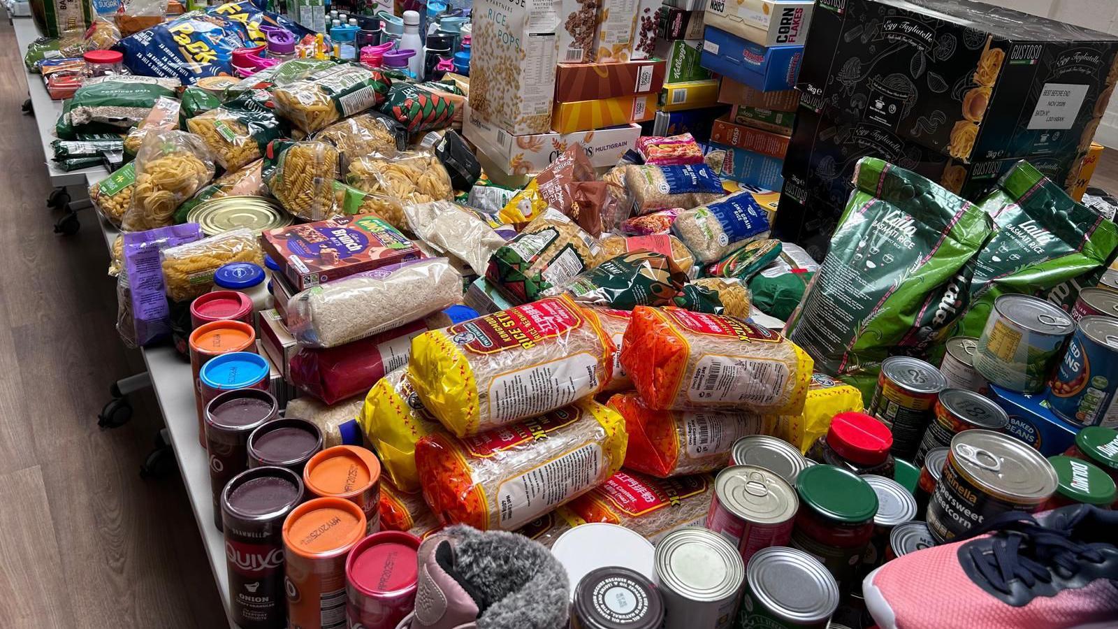 Two tables full of donations including tins of food, packets of pasta and pink trainers