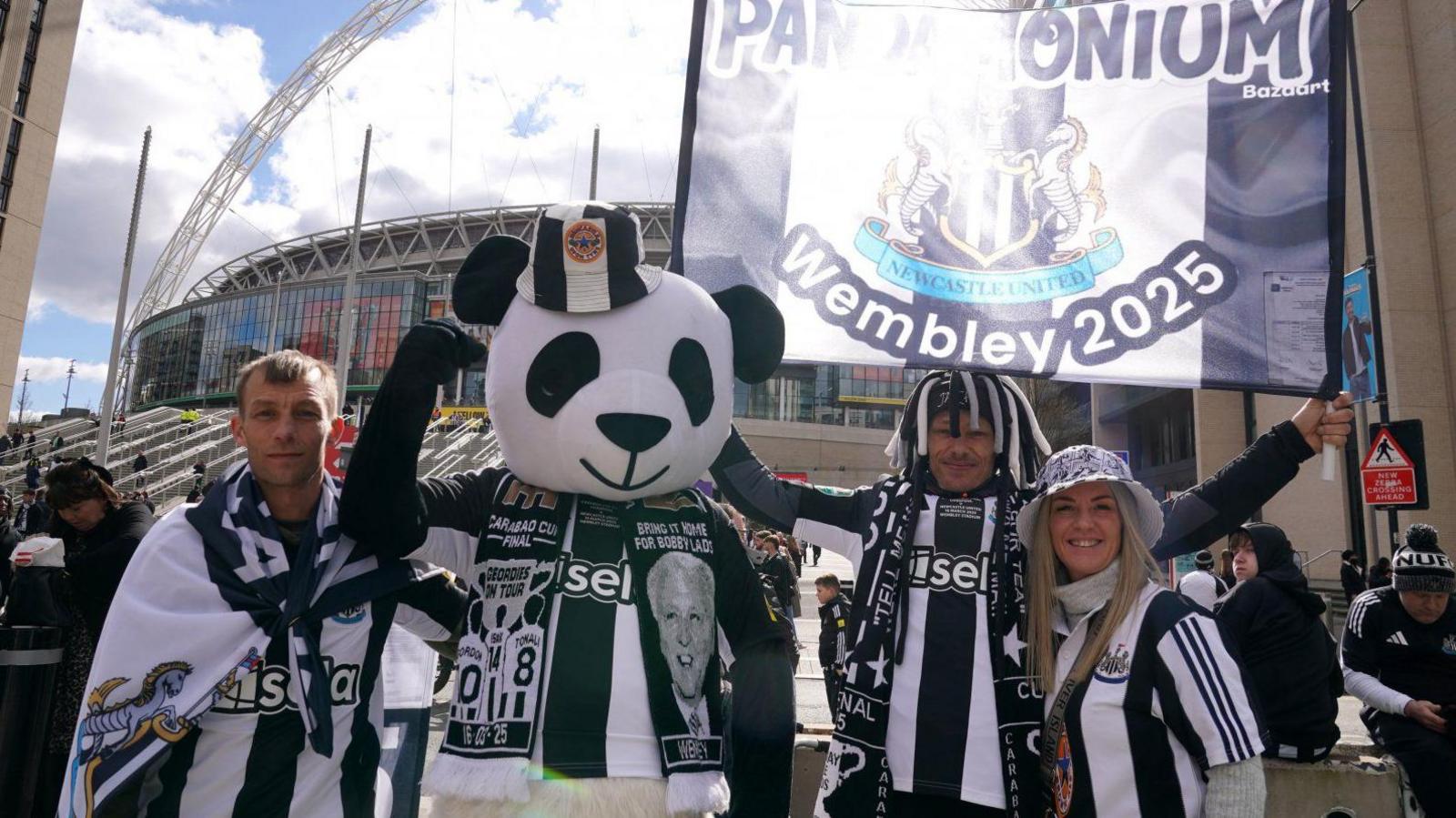 Two men and a women in Newcastle United strips with another person dressed as a giant panda. They are holding aloft a banner and Wembley Stadium can be seen in the background