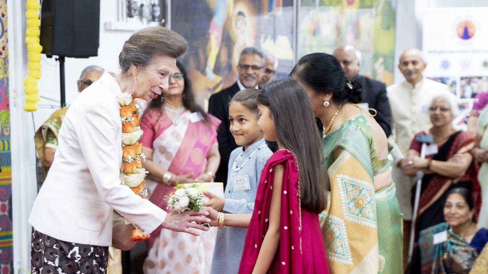 A woman in a white top in an orange garland shakes the hands of three women in Hindu dress.