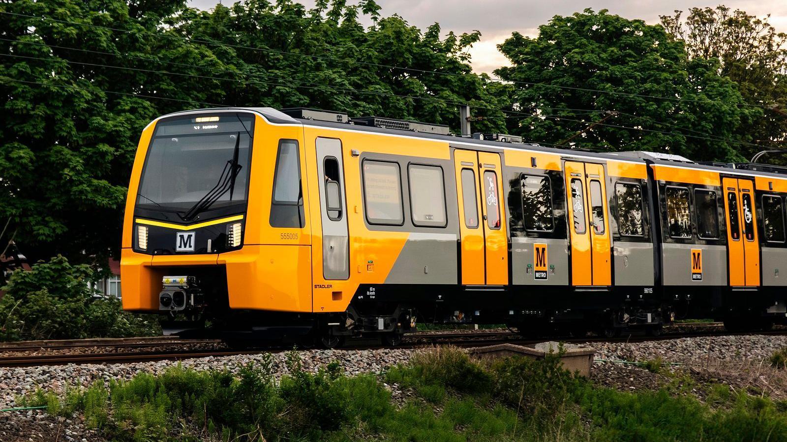 One of the new Metro trains, with bright yellow and some grey colouring. It is travelling on a train track. There is small green shrubbery in the foreground of the picture and also some green trees in the background behind the train too.