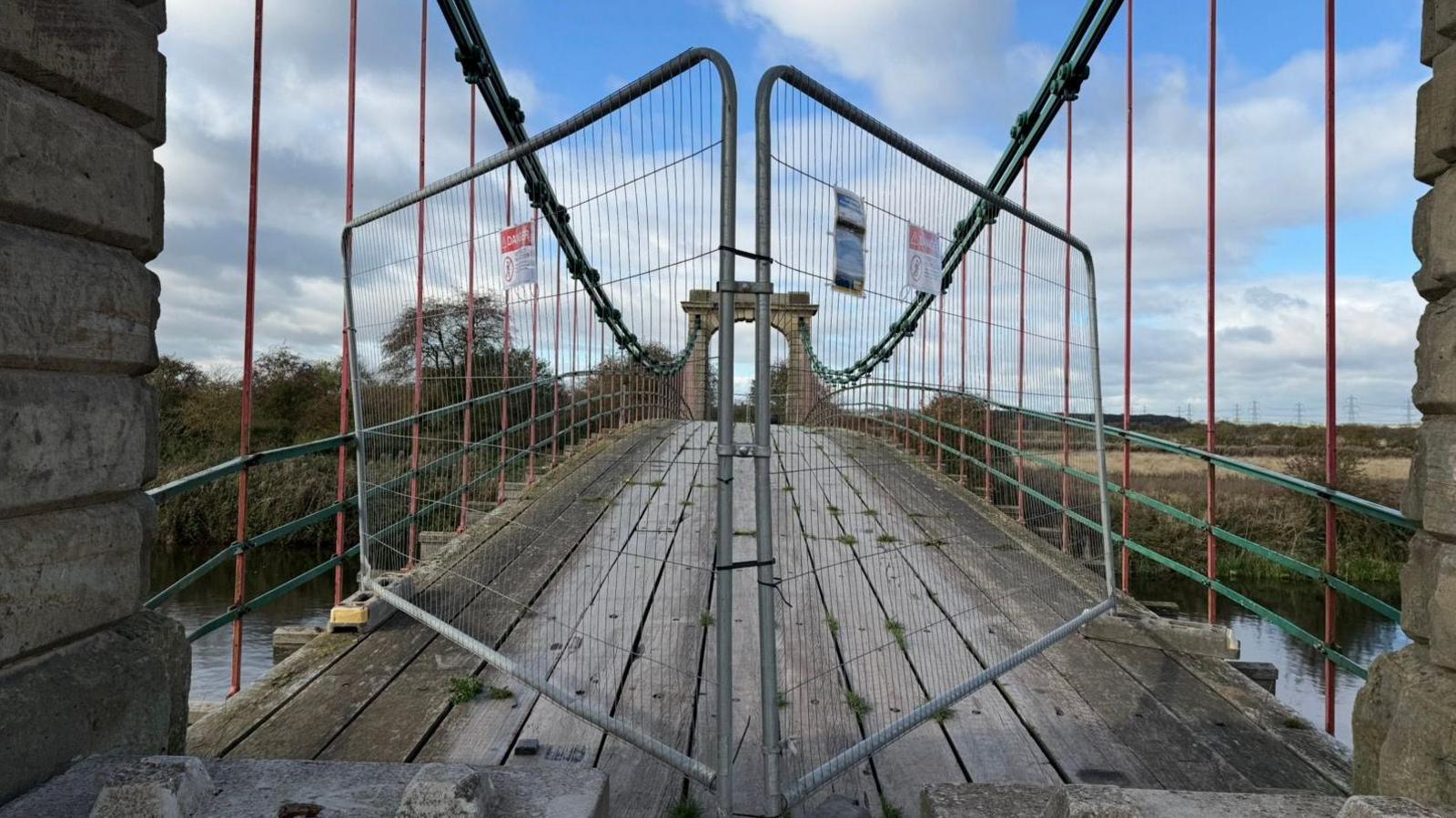 The picture has been taken from one side of an old style suspension bridge. The floor is wooden, and curves in the distance as it reaches the other side. There are red and green metal bars either side of the bridge, preventing it from being open at each side. Directly in front of the camera are two metal fences, preventing people from passing. One of the signs on them reads 'Danger.'