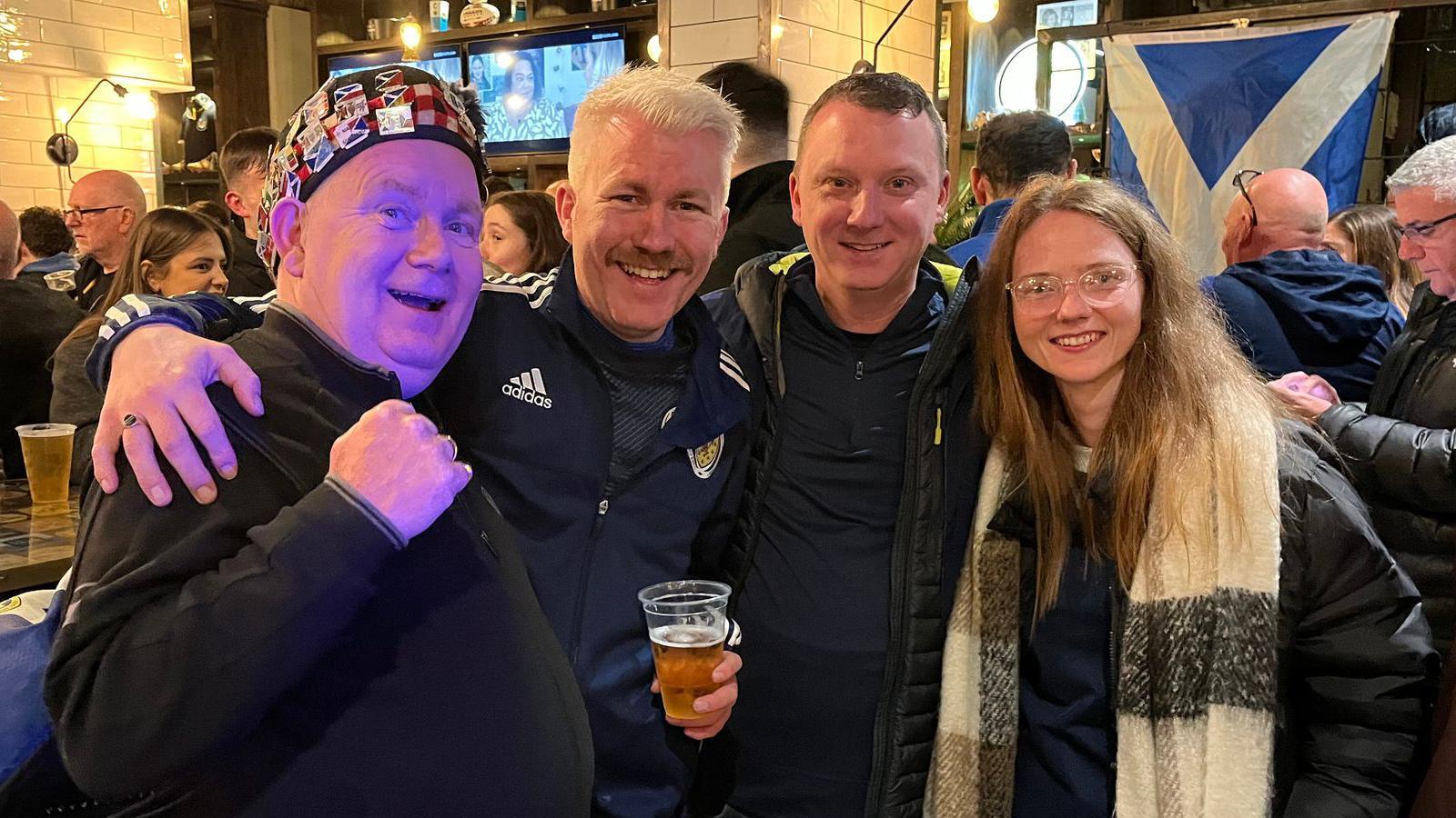Three men and a woman stand smiling with their arms around each other in a crowded pub. A saltire hangs in the background and one of the men holds a beer