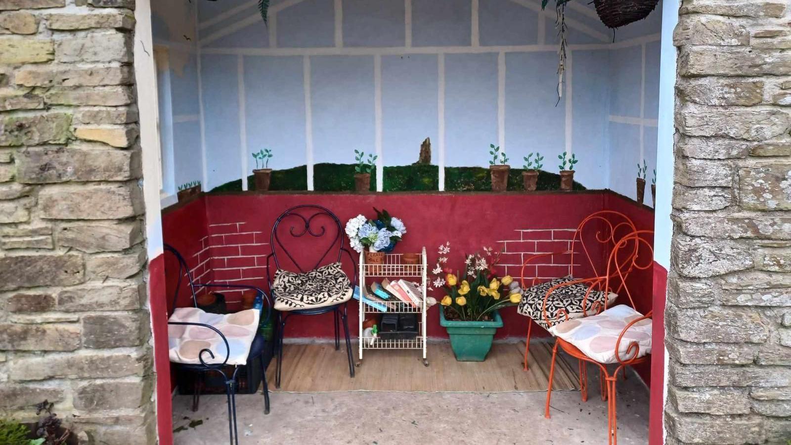 The inside of a stone bus stop which has been painted. There bottom half of the bus stop has been painted dark red to look like bricks. The top half has several painted plant pots in front of a blue sky background that looks like it's inside a greenhouse. There are four metal chairs inside the bus stop with cushions on. There is also a small book shelf with a flower pot on the top level, and a green plant pot full of tulips on the floor beside it.